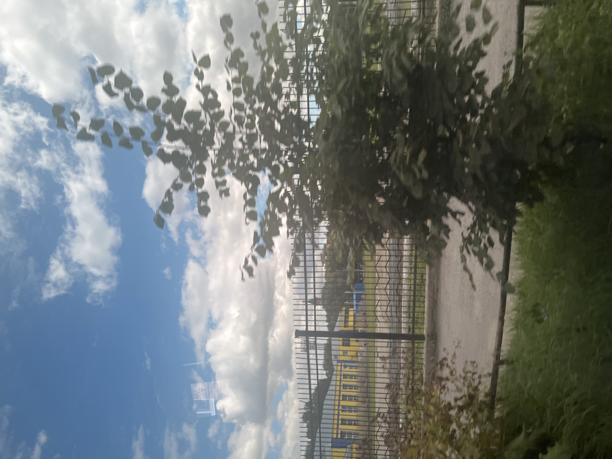 The image depicts a view through a window, likely from a moving vehicle, in Ulyanovsk, Russia. In the foreground, a tree with blurred leaves partially obscures the view. Behind the tree, a black metal fence runs horizontally across the image. Beyond the fence, a yellow building with a blue roof is visible, featuring multiple windows. In the distance, a dark tower with a red top stands against a sky filled with white clouds and blue patches. The ground in front of the fence appears to be a path or road, and to the left of the fence, green grass and foliage are present. The overall scene suggests an urban or suburban environment on a cloudy day.