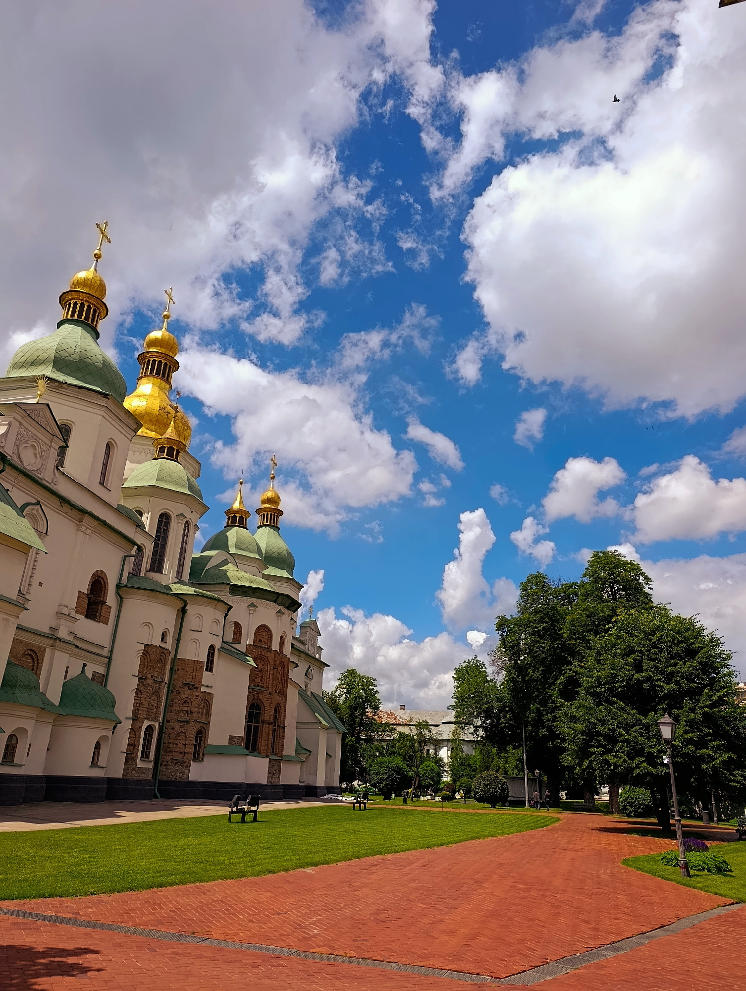 The image depicts Saint Sophia's Cathedral in Kyiv, Ukraine, on a clear day with scattered white clouds against a bright blue sky. The cathedral's white facade is adorned with green domes topped with golden crosses. In the foreground, a brick pathway leads into a manicured green lawn. Several benches are visible on the lawn, and a lamppost stands near a large, leafy tree. In the distance, other buildings and trees can be seen. A single bird is visible flying in the sky.