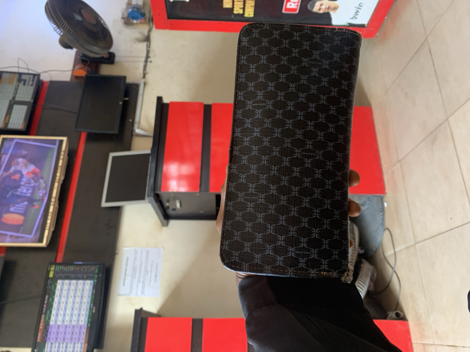 A person wearing a black long-sleeved shirt is holding a dark-colored wallet with a repetitive pattern in a room that appears to be a betting shop. The location is possibly Adankolo, Nigeria. There are multiple screens displaying data, including one showing what appears to be sports-related content. The room has red accents on the furniture and walls. There's an electric fan mounted high on a wall, and a document is posted near one of the monitors. The wallet's surface appears patterned with an abstract design of repeating elements.