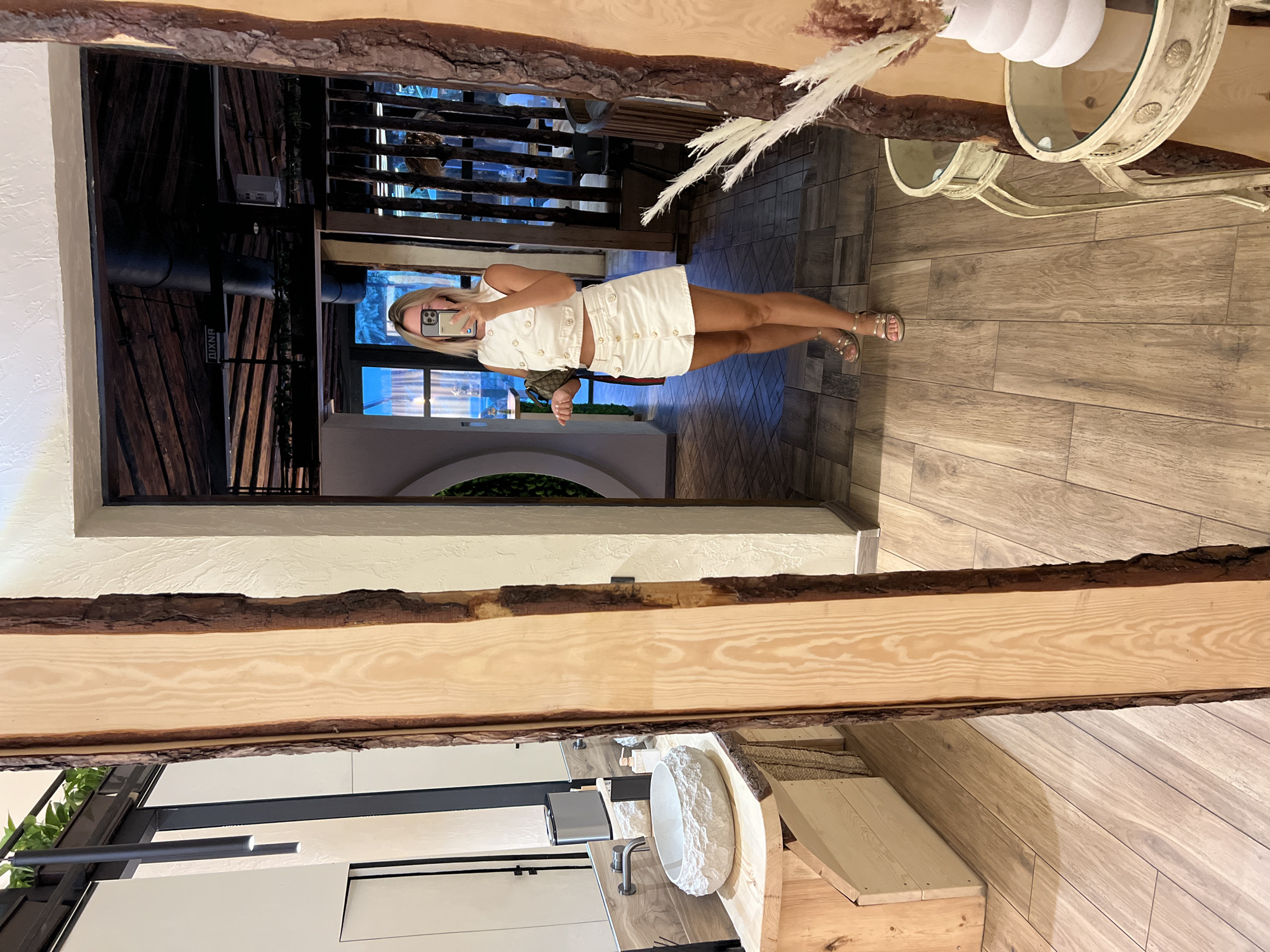 A blonde woman in a white skirt and top takes a selfie in a rustic-themed interior. She is standing in front of a large mirror framed with rough-hewn wood. The mirror reflects the interior of what appears to be a restaurant or lounge area. The woman is wearing gold sandals and carrying a small purse. In the reflection, we see wooden beams and tables, as well as other patrons in the background. To the left of the woman, in the foreground, there is a bathroom vanity with a stone sink. The floor is tiled with wooden planks. The scene is dimly lit, with warm light casting shadows. The location is identified as Odesa, Ukraine.