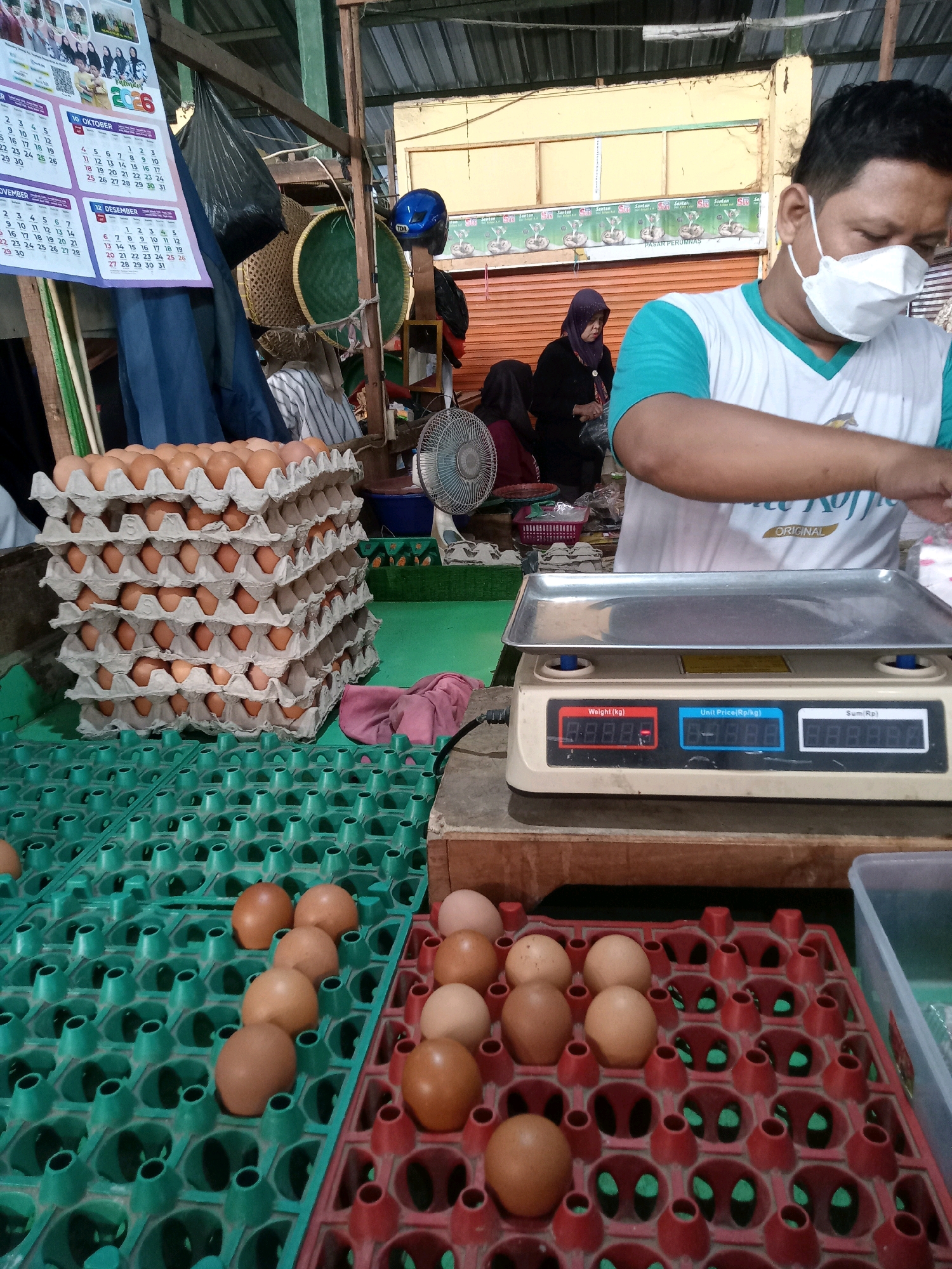 The image shows a market stall in Cirebon, Indonesia, with a large stack of egg cartons filled with brown eggs. A vendor, wearing a face mask and a blue and white shirt, is standing behind a digital scale and appears to be preparing to weigh or package the eggs. In the background, two women are visible, one wearing a purple hijab. Various market items, including a calendar, bags, and other egg cartons, are also present. The environment suggests an indoor market setting with a metal roof.
