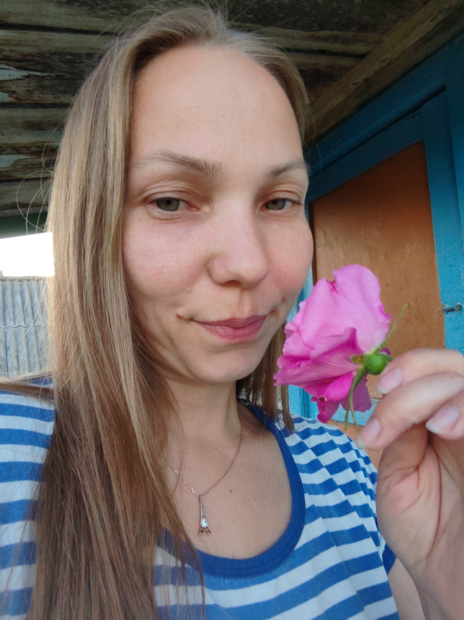 A medium shot from a slightly low angle shows a blonde woman with light brown hair wearing a blue and white striped t-shirt, holding a pink rose in her right hand. She is looking forward and slightly to her left with a subtle smile. The background consists of a wooden roof, a blue painted door, and a corrugated metal fence. The scene appears to be outdoors, possibly in a rural or suburban setting in the city of Chuchlem, Russia, suggested by the overall aesthetic of the environment. The woman's necklace, a thin gold chain with a small diamond pendant in the shape of the Eiffel Tower, is visible. The lighting is natural and appears to be daylight.