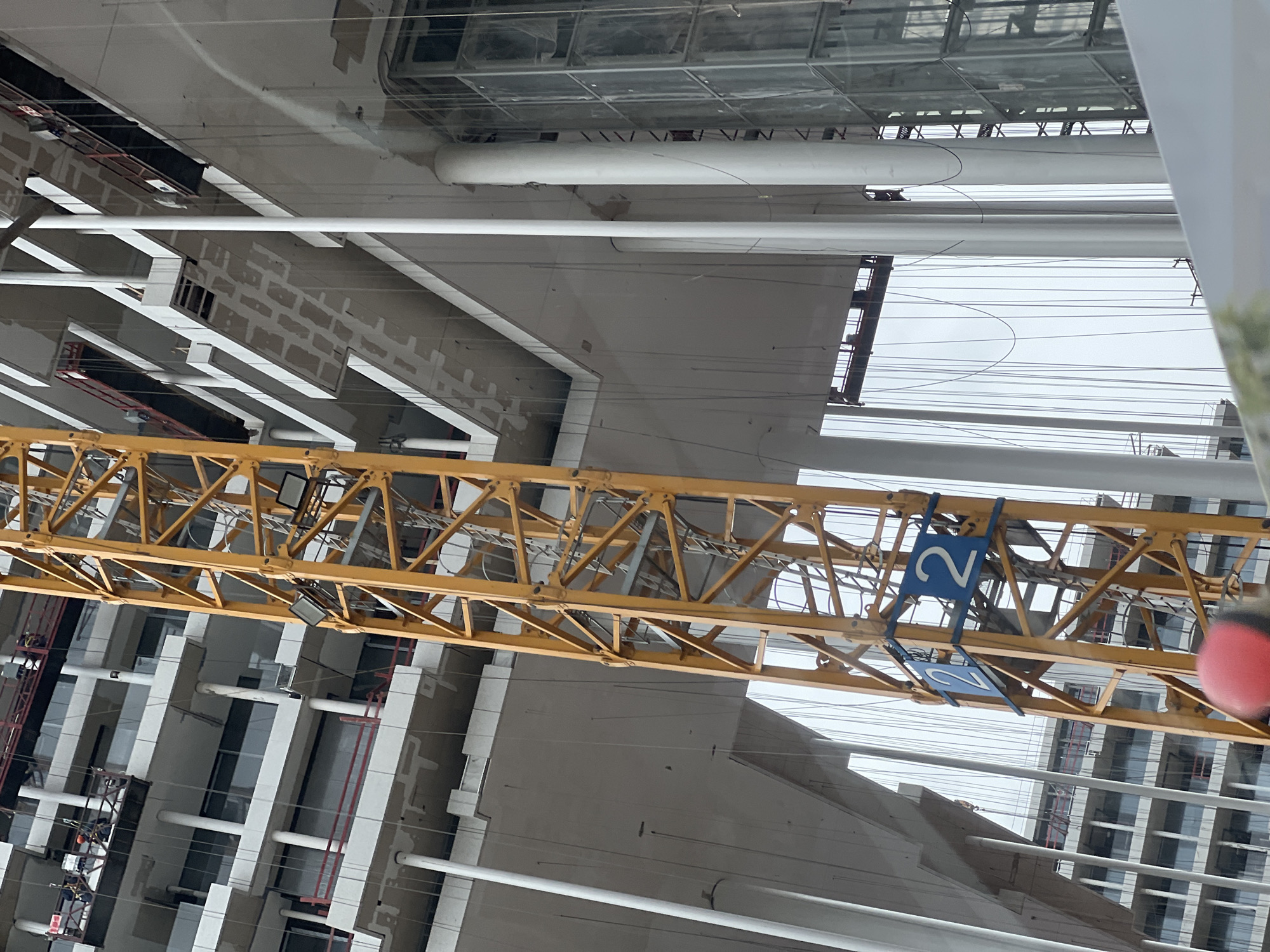 The image depicts a construction site in Khimki, Russia. A yellow crane, marked with the number "2", dominates the scene. Buildings under construction are visible, featuring numerous windows and white structural elements. Several white pipes and numerous thin wires crisscross the sky. There is no visible human presence. The sky is overcast.