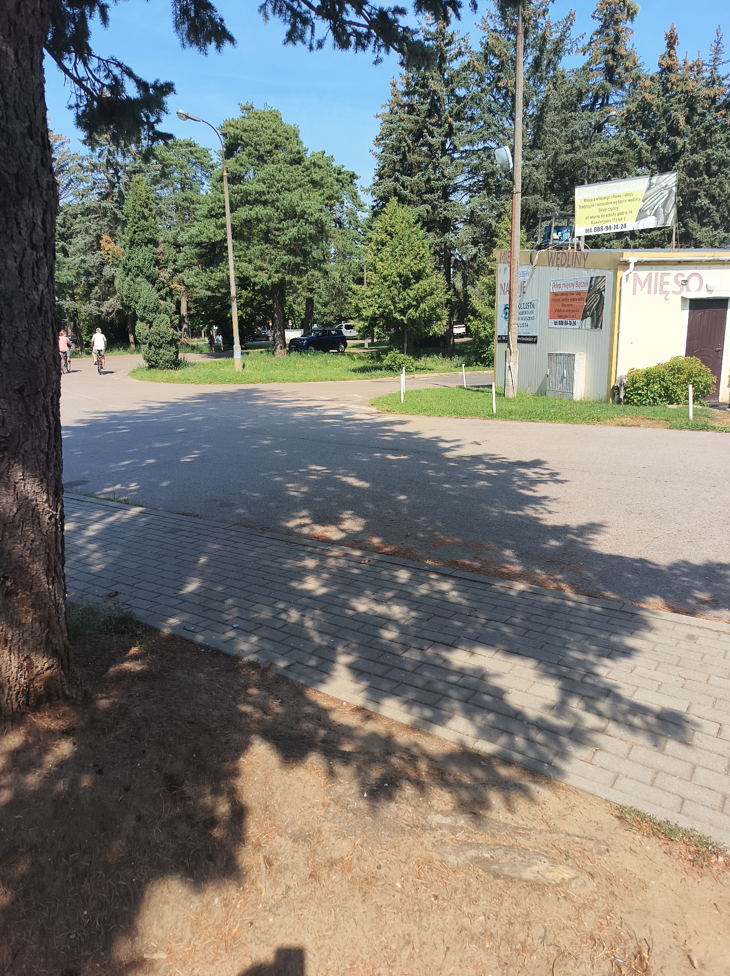 In Białystok, Poland, on a sunny day, a paved pathway with a gravel edge borders a grey asphalt road. Two individuals on bicycles ride away from the camera on the road, which is lined with trees and grass. A light post stands near a building with a sign advertising "Mieso Baczek" (Meat Shop). The building has a yellow facade and a brown door. Shadows of tree leaves are cast across the pathway and the road.