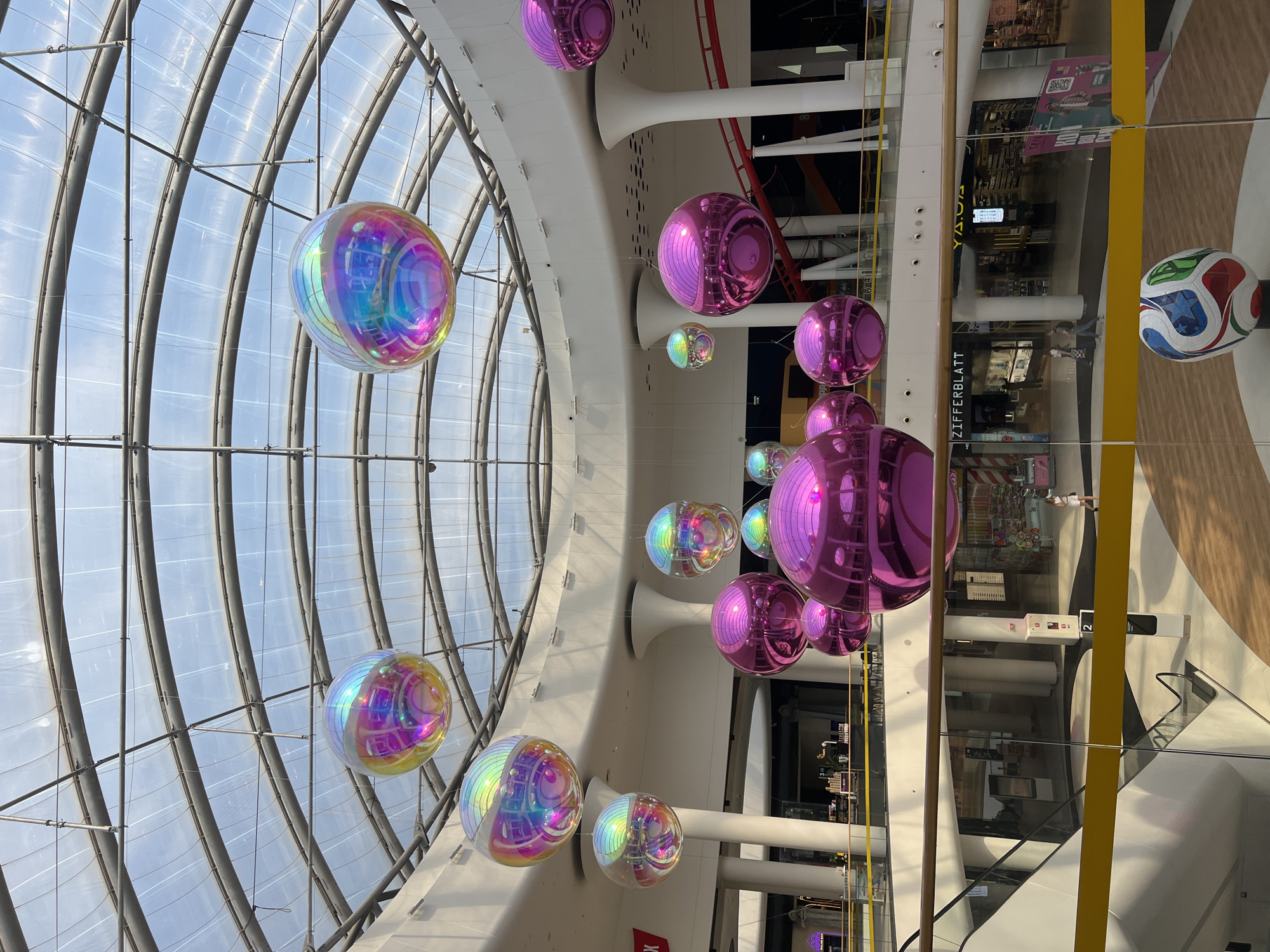 The image shows the interior of a modern shopping mall with a high, domed glass ceiling. Suspended from the ceiling are numerous large, spherical ornaments, some with iridescent, multicolored surfaces, and others with a shiny pink finish. The mall features multiple levels, escalators, railings, and storefronts visible in the background. A large, decorative soccer ball sculpture is positioned on the ground floor in the foreground. The scene is brightly lit by natural light filtering through the ceiling and artificial mall lighting. No specific landmarks or indicators of the city of Kyiv, Ukraine are visible in the image.