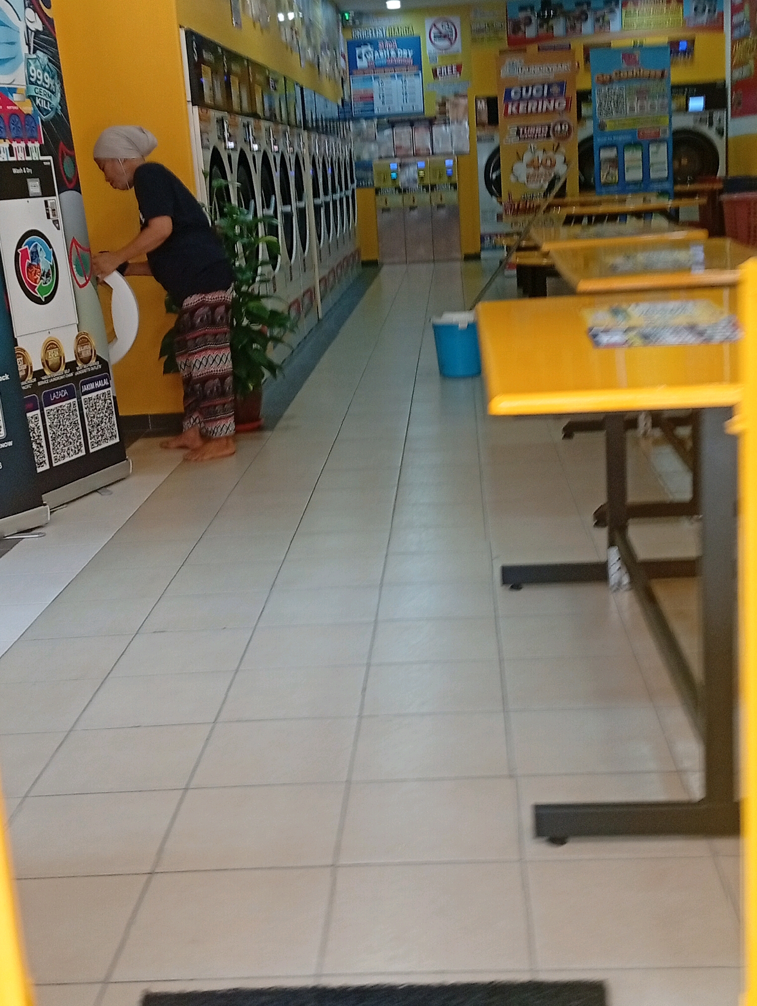 A full-shot, eye-level, slightly blurry, indoor image shows a laundromat. In the foreground, a person wearing a headscarf and patterned pants is operating a washing machine. The person is barefoot. Behind the person, a row of washing machines extends into the background. Various signs are visible on the walls, some with text in Malay, advertising laundry services and promotions. Yellow tables and chairs are set up in the mid-ground, suggesting a waiting area. A blue bucket is visible on the floor near the tables. The floor is tiled in a light color. The overall scene depicts a public laundromat in Selayang Municipal Council, Malaysia, with a customer engaging in laundry services.