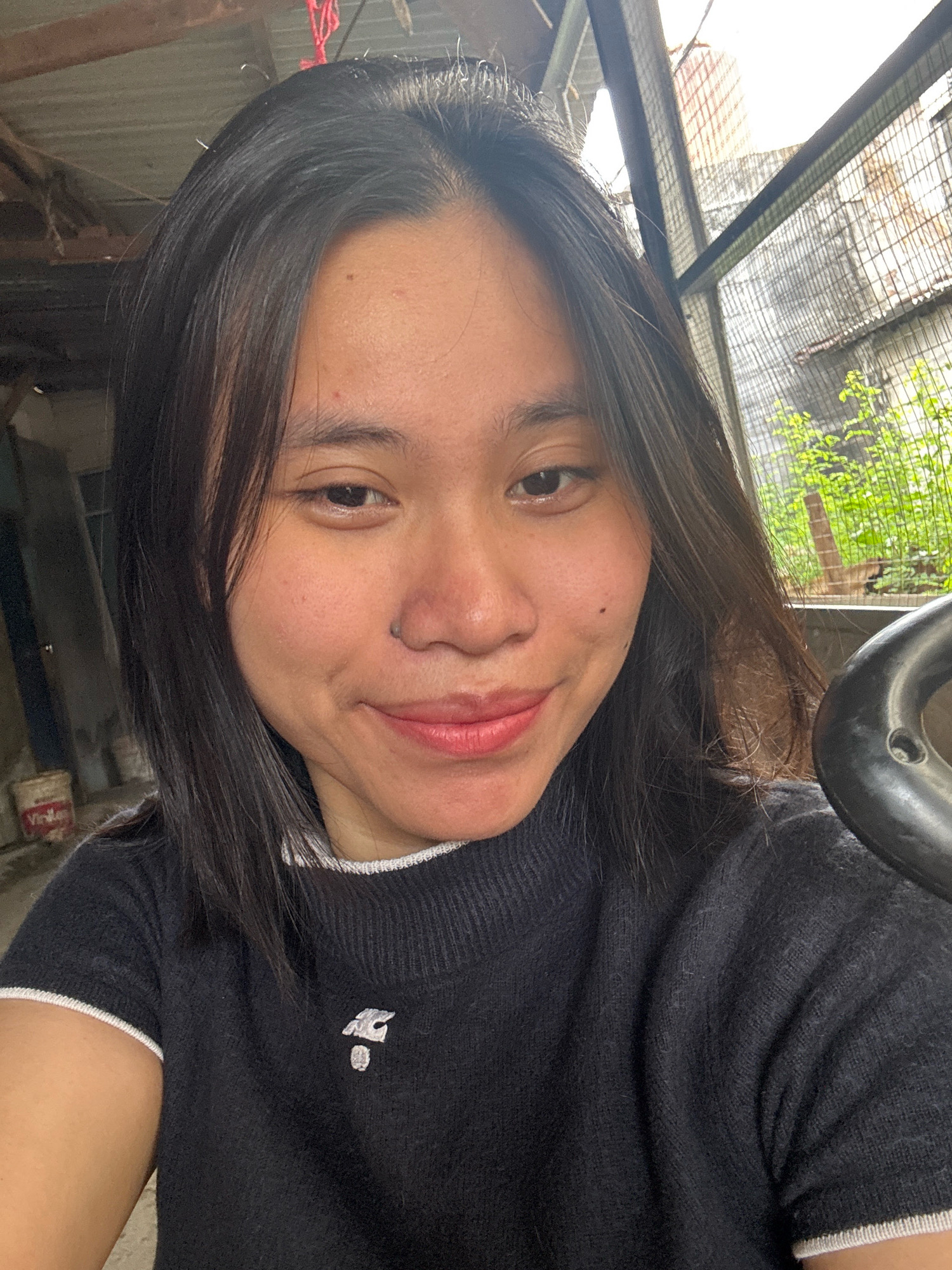 A medium shot, selfie-style photograph shows a young East Asian woman from the chest up. She has long, dark brown hair that falls past her shoulders. She is wearing a black, short-sleeved sweater with a white trim at the collar and sleeves. A small, silver emblem is affixed to the left side of the sweater's chest. Her skin is smooth with a few small blemishes on her cheeks. She has a mole on her right cheekbone and a slightly darker mole below her left nostril. Her lips are painted a soft pink. The background suggests an outdoor or semi-outdoor setting. To the left, there is a textured wall and a portion of a bucket with red and white writing. Behind her, a metal fence encloses an area with lush green vegetation. Above the fence, a slightly dilapidated building with a corrugated metal roof is visible. A thick, dark, curved object, possibly a railing or pipe, is on the right side of the frame, partially obscuring the woman's right shoulder. The lighting is bright, indicating daylight. The location is identified as East Jakarta, Indonesia.