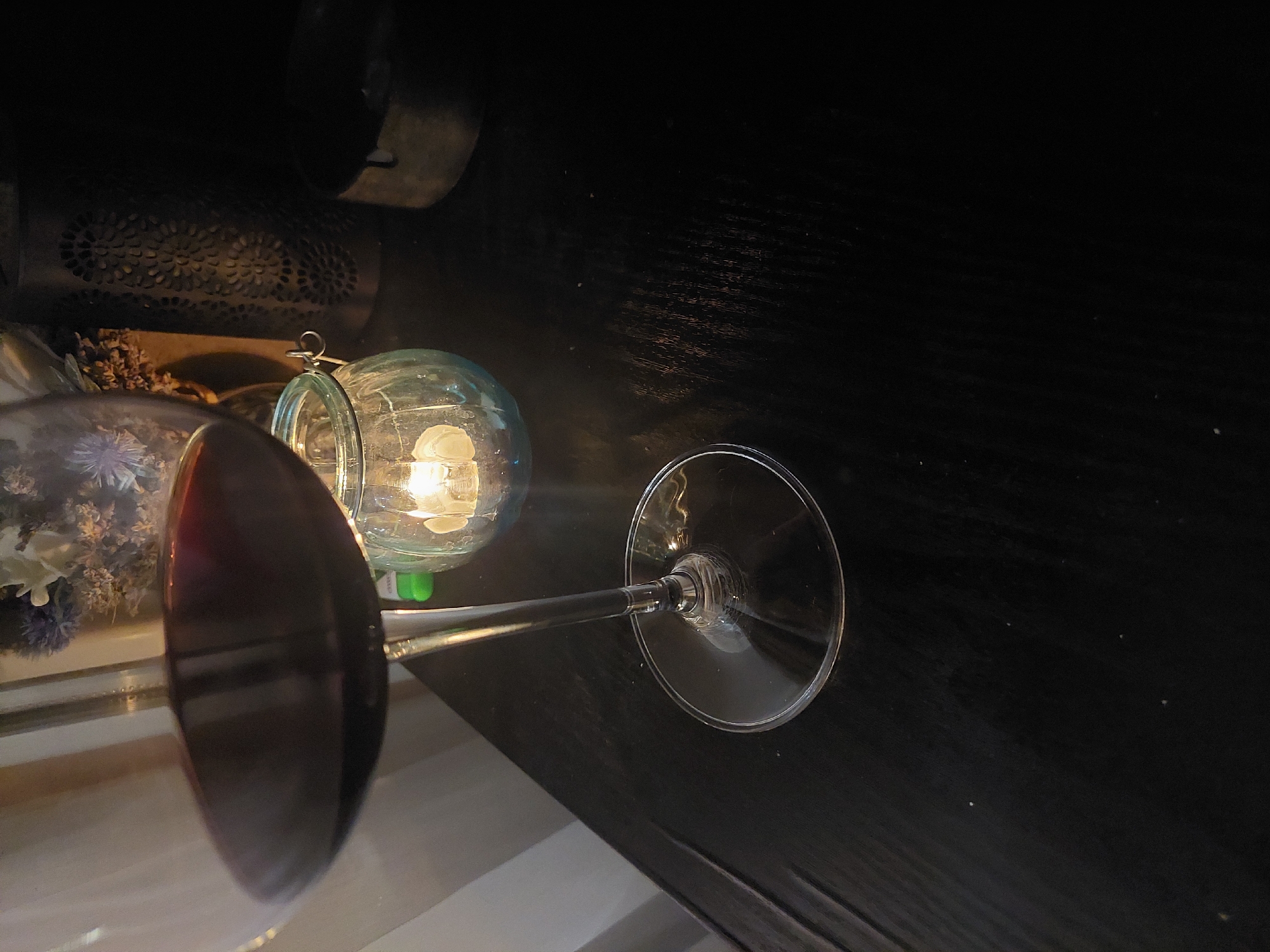 The image shows a dimly lit interior scene. A wine glass, nearly full of a dark red liquid, dominates the foreground. The glass is situated to the left of the frame. To the upper left and somewhat behind the wine glass is a clear, glass jar containing a lightbulb, which is illuminated. Behind the jar and further back is an object that appears to be a metal container with a decorative, patterned surface. The rest of the background is a dark, reflective surface, possibly a table.
There is no indication in the image regarding the city of Plovdiv, Bulgaria.
