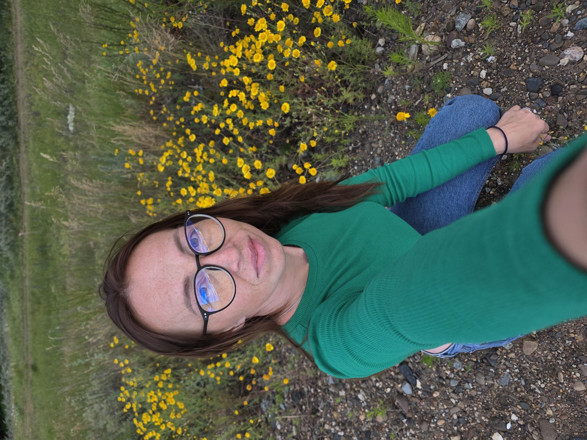 A woman with brown hair and glasses kneels in a field of yellow flowers and dry grass. She is wearing a green long-sleeved shirt and blue jeans. The ground is covered in gravel and small rocks. The background consists of a green hill or treeline. The image appears to have been taken in a rural or natural setting, possibly on the outskirts of Chaikovsky, Russia, given the presence of wildflowers and natural vegetation characteristic of the region.