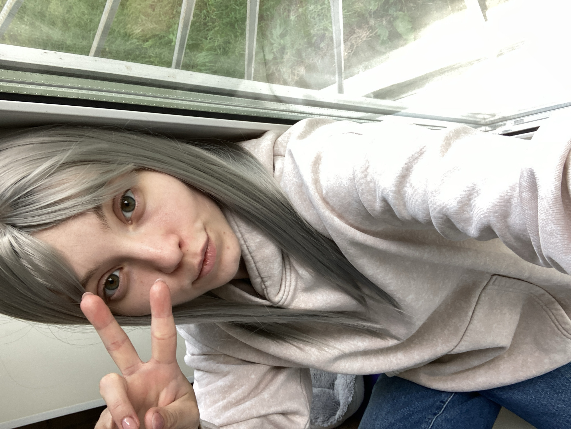 A young woman with light skin, green eyes, and long, straight, grey hair is taking a selfie. She is wearing a light pink hooded sweatshirt and blue jeans. She is holding up her left hand with two fingers extended in a peace sign. She is sitting next to a window, and through the window, there is a view of green grass and trees. The lighting is bright, suggesting it is daytime. The image quality is clear. The overall scene is casual and relaxed.