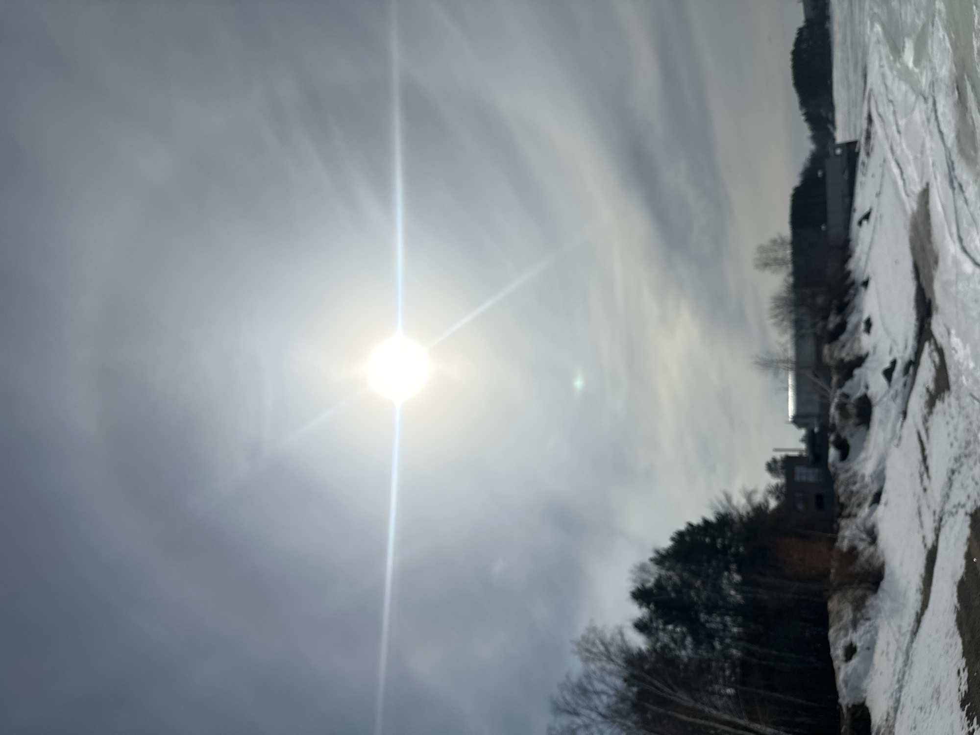 The image depicts a winter scene in Chaikovsky, Russia, with a bright sun partially obscured by thin clouds, causing a halo effect. A distinct vertical light pillar extends from the sun. The foreground shows a snow-covered bank with a mixture of bare trees and evergreens. Buildings, including what appears to be residential structures, are visible among the trees. A frozen body of water, possibly the Kama River, is partially visible with ice formations along the shoreline. The sky is overcast with diffused light. No people are discernible in the image.