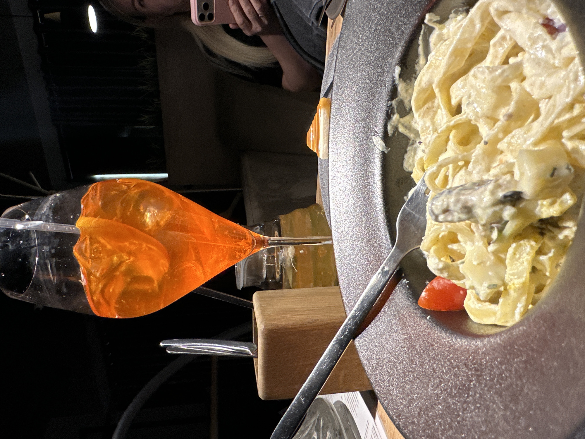 A person holds a pink mobile phone above a table in a dimly lit setting. On the table, there is a glass with an orange liquid inside, a wooden stand holding cutlery, and a metallic bowl filled with pasta covered in a creamy sauce with a single tomato piece on the side. The location appears to be in Chaikovsky, Russia.