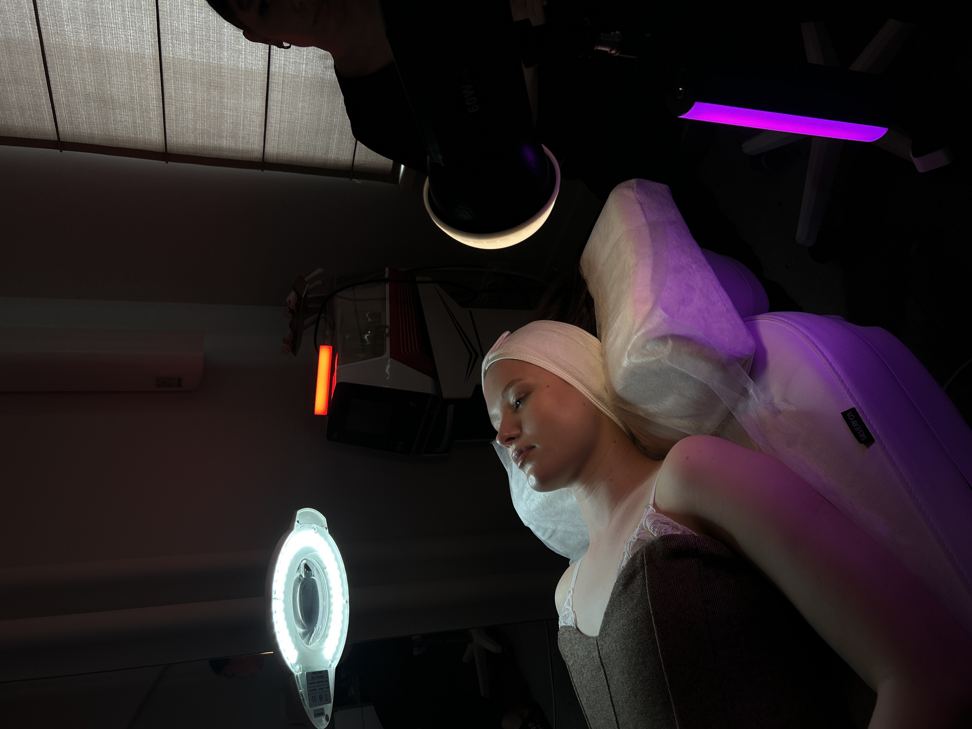 A person is positioned in a cosmetology chair with a white head covering. The lighting is a combination of ring light, overhead fluorescent, and a directional light. The chair is branded Silverfox. A person is partially visible in the upper portion of the frame. The location cannot be verified as Izhevsk, Russia, without additional information.