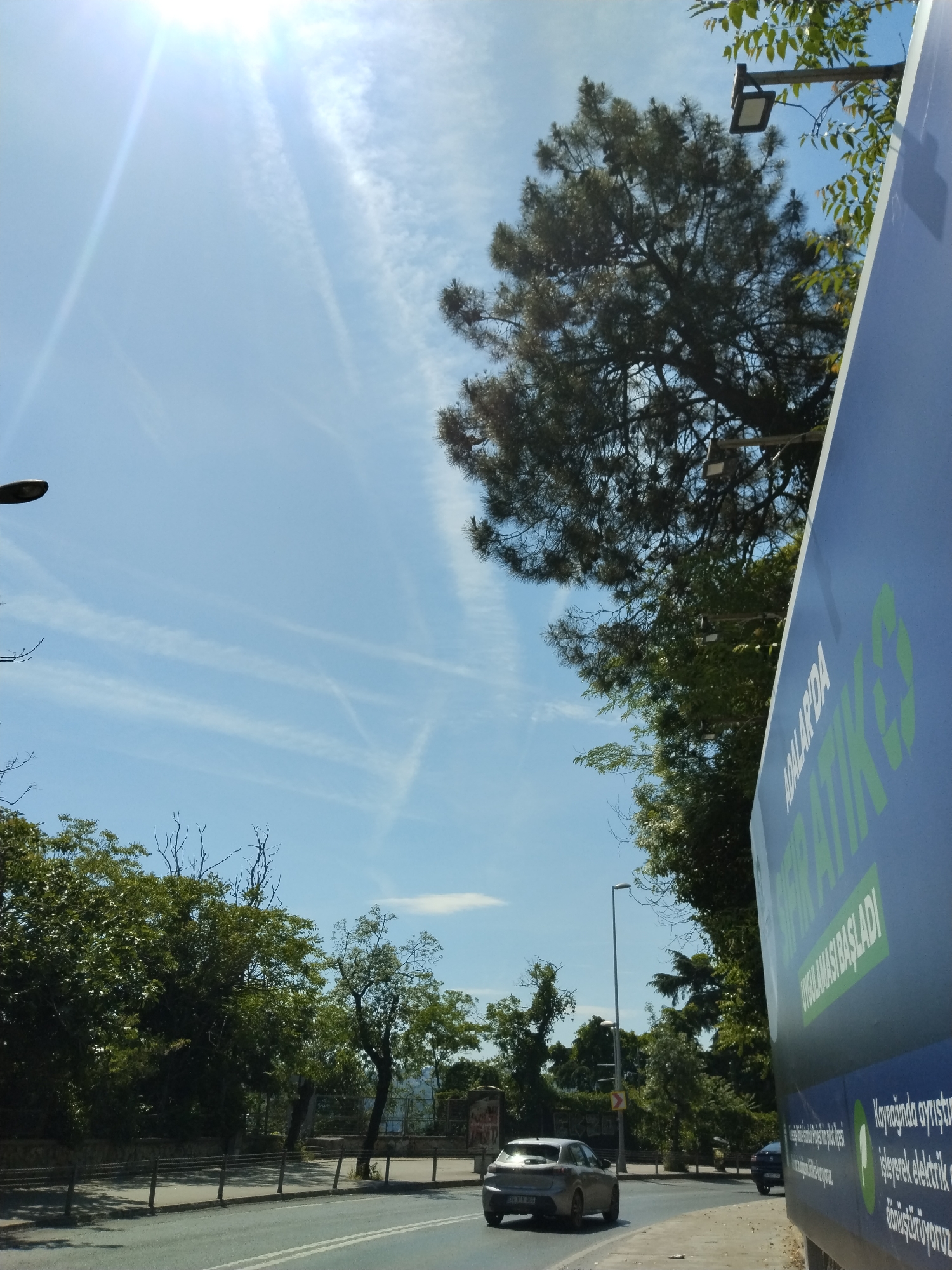 This image depicts a sunny outdoor scene on a road in Sarıyer, Turkey, likely during midday or early afternoon. The sky is a bright, clear blue, dominated by a strong sun in the upper left that creates prominent lens flare. Several white contrails from aircraft are visible, crisscrossing the sky.
On the right side of the frame, a large, blue billboard is angled towards the viewer, partially revealing its text and a dense pine tree behind it. The billboard, adorned with a green recycling symbol, displays Turkish text in white and green. Visible phrases include "ADALAR'DA" (In the Islands), "AMBALAJ ATIĞI TOPLANIYOR" (Packaging Waste Is Being Collected), and "UYGULAMASI BAŞLADI" (Application Started). Smaller text at the bottom describes the process: "Kaynağında ayrıştırıp işleyerek elektrik üretiyoruz / dönüştürüyoruz," which translates to "Separating at its source, we produce/recycle electricity by processing it."
The road, appearing to be asphalt with white double lines, stretches into the distance. A silver hatchback car, with a partially visible license plate "34 B... 389" (indicating Istanbul), is driving away from the viewer on the left side of the road. A darker vehicle is also visible further ahead on the right. Both sides of the road are flanked by abundant green foliage and various trees, some with thick canopies and others with more visible branches. Several modern streetlights with square LED fixtures are interspersed among the trees. The overall impression is that of a typical, busy street on a clear and bright day.