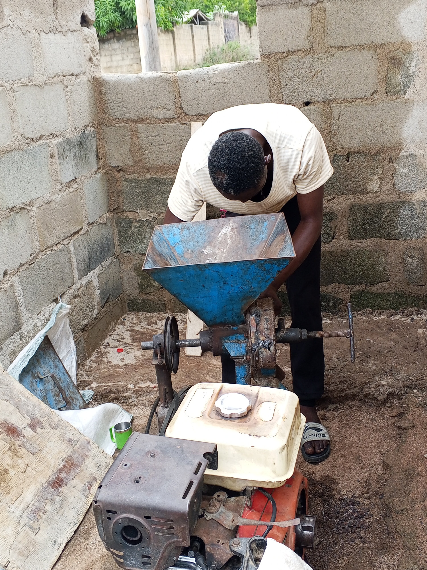 An individual in Zango-ndaji, Nigeria, is operating a machine, possibly a grain mill, located in what appears to be an unfinished structure with exposed concrete blocks. The person is wearing a striped shirt, black pants, and sandals. The machine consists of a blue hopper above a dark, engine-like base with a white fuel tank. A wheel and a handle are visible on the machine's side. A green mug, some wood, and a white bag are on the ground near the machine. The ground is dirt.