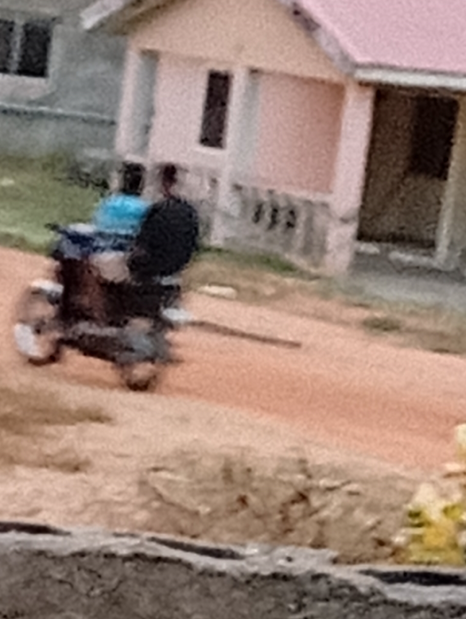 A grainy photograph depicts a blurry scene with a residence in the background and a vehicle in the foreground. The residence is a single-story structure with a pink roof. A two-wheeled vehicle is in the middle of the scene. Two people are on the vehicle. The scene appears to be a street in a residential area with dirt ground. The location is Unknown, Unknown.