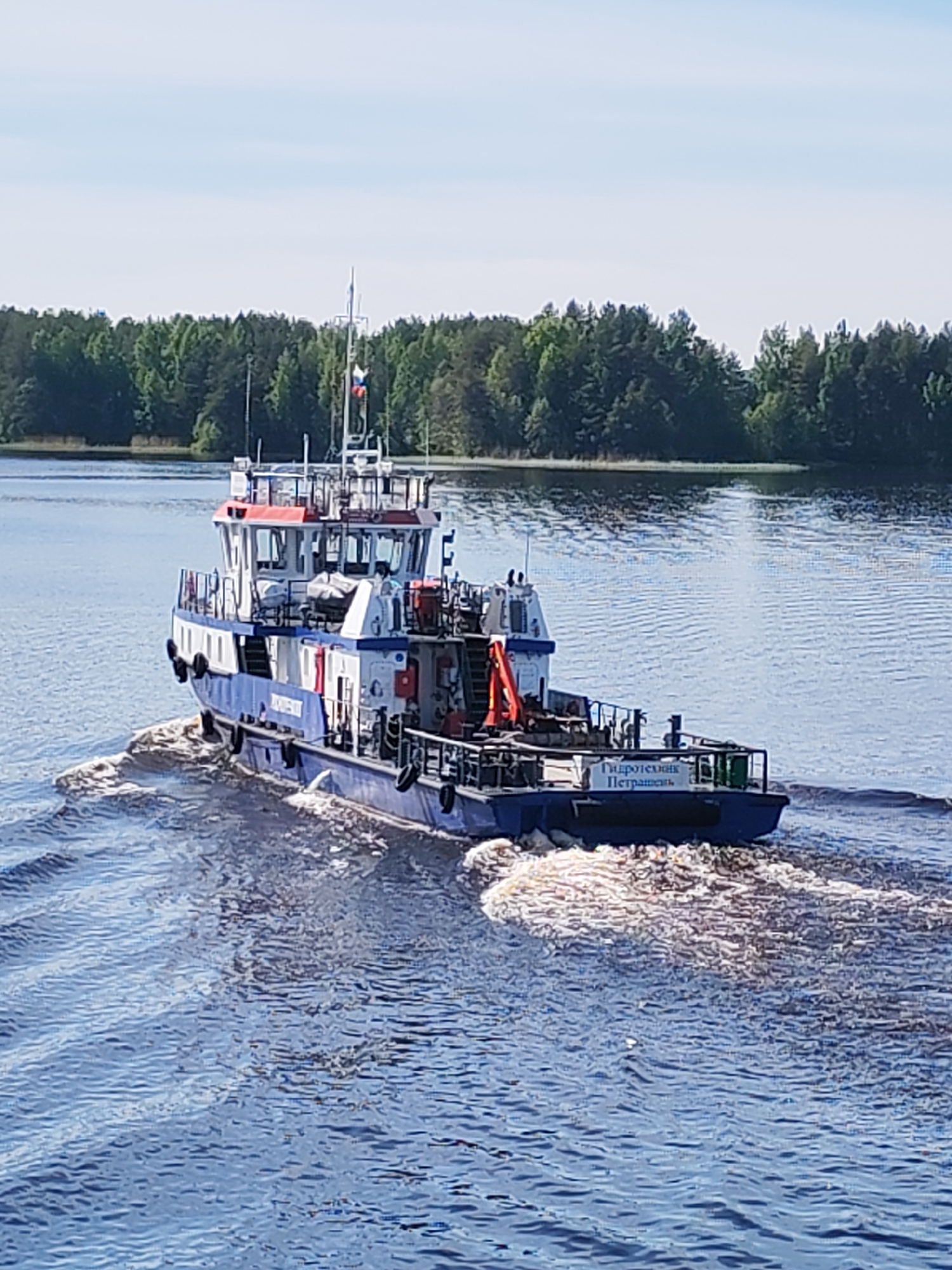 An aerial-level shot captures a blue and white workboat, possibly a tugboat, moving across a wide body of water, leaving a pronounced wake behind it. The boat, seen from its stern-port side, features a blue hull and a white superstructure with red accents on the cabin roof. A Russian flag is visible flying from a mast at the top of the cabin. On the boat's deck, an orange crane is prominent, along with various other equipment and railings. Black fenders are attached to the hull's sides.
The water is calm with ripples and the churned wake from the boat's propulsion. In the background, a dense line of green coniferous trees forms the distant shoreline under a clear, light blue sky, suggesting a bright sunny day.
Visible text on the port side of the hull, towards the bow, reads "ИНЖЕНЕР" (INZHENER). On a white panel at the stern, the text "Гидротехник Петрашень" (Gidrotekhnik Petrashen') is clearly legible. The scene is set in Ozerki, Russia, likely on a lake or a wide river.