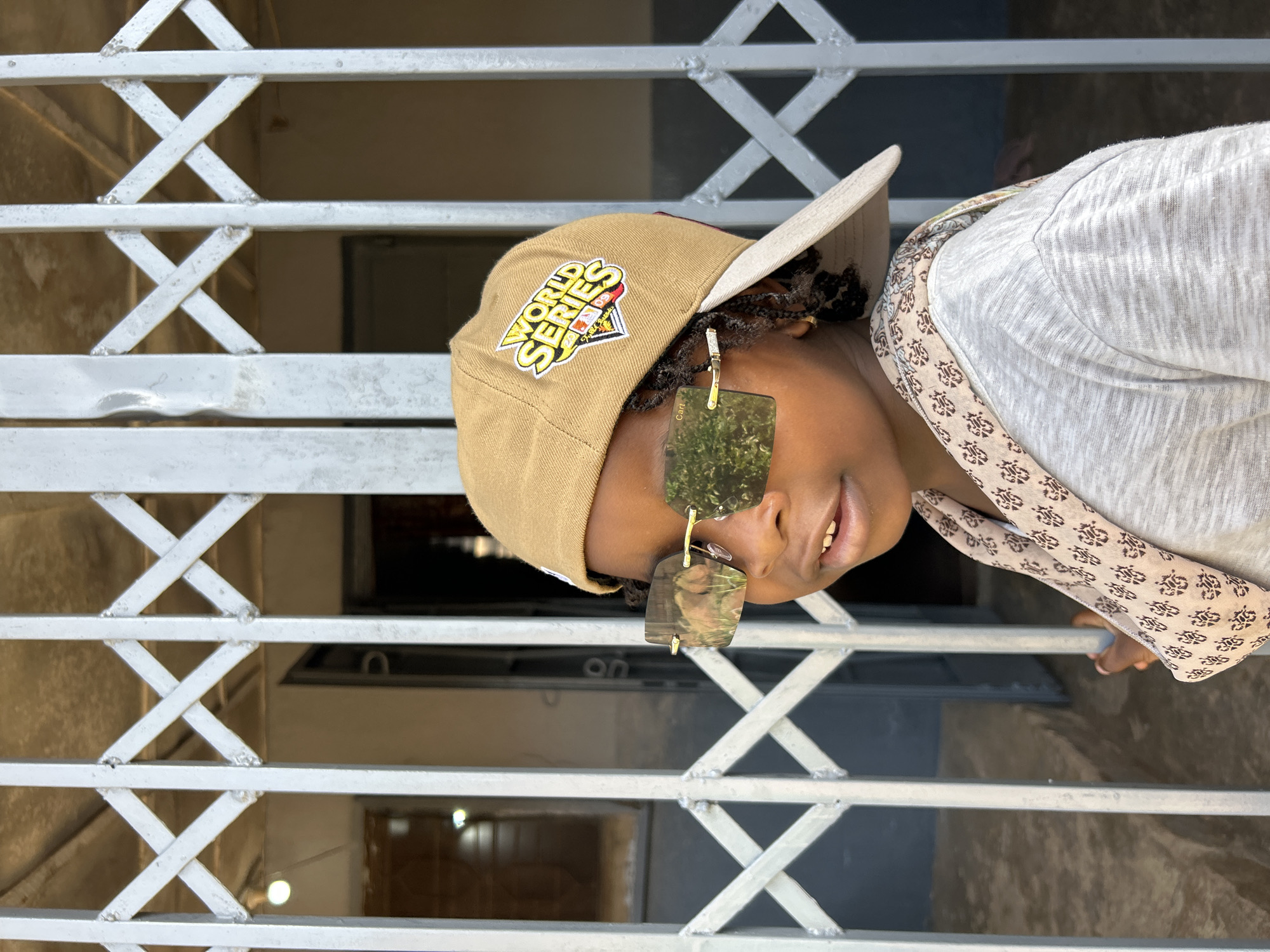 A person wearing a khaki baseball cap, a light-colored patterned shirt, and dark-tinted glasses stands behind a gray metal security gate. The hat has a "World Series" logo on the front. The person is smiling slightly. The gate has a criss-cross pattern and appears to be in front of a building with a doorway visible in the background. The scene is likely in Jos, Nigeria.