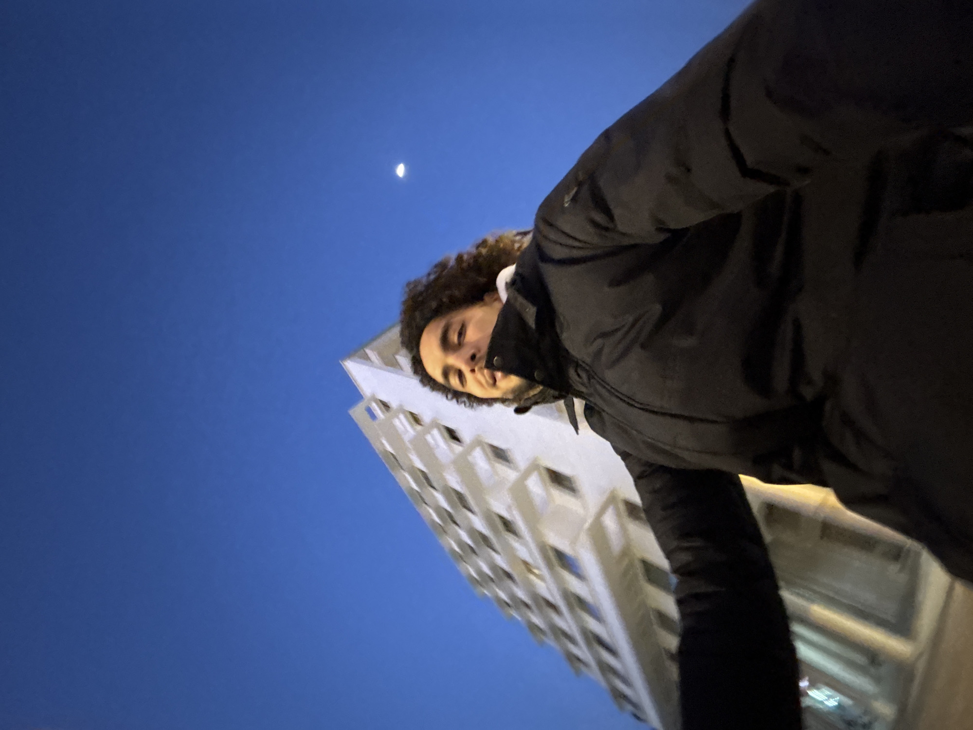 A low-angle shot shows a person in a black winter jacket, with curly hair, standing outside at night. The person is partially obscured by the jacket, but their face is visible. Behind the person is a tall, white building with many windows, suggesting an apartment complex or office building. In the upper left corner of the frame is a small crescent moon in a deep blue sky. The photo is taken from a skewed perspective, likely a selfie. Based on the architecture, the location is likely Tanger (Tangier), Morocco.