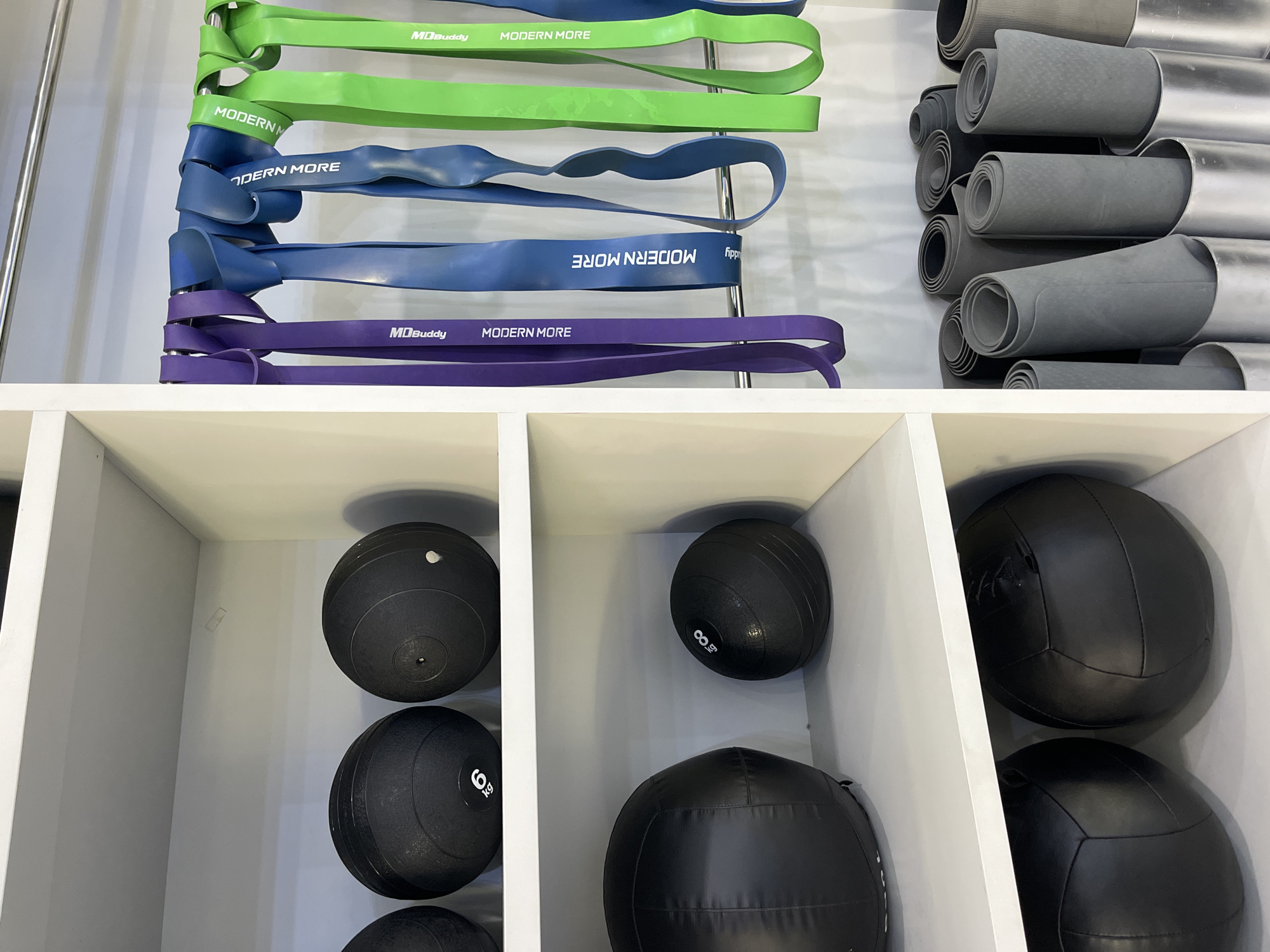 The image depicts a storage area within a gym located in North Kuta, Indonesia. On the left side of the frame, three shelves are filled with various fitness equipment. The top shelf displays two black medicine balls, one of which has a white marking indicating "6 kg". The middle shelf features two larger black medicine balls, one significantly larger than the other. The bottom shelf holds two more black medicine balls. To the right of the shelves, a metal rack is adorned with a collection of resistance bands in purple, blue, and green. Further to the right, several rolled-up yoga mats are neatly stored in cylindrical containers. The environment appears clean and organized, suggesting a well-maintained fitness facility.
