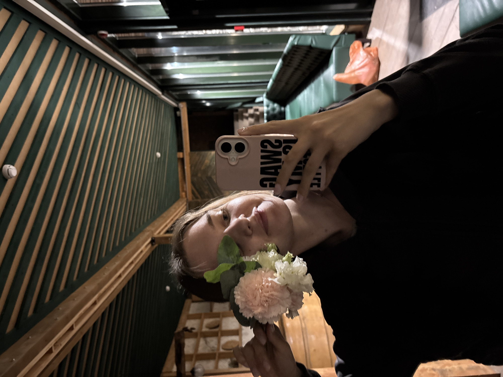 A medium shot, eye-level selfie captures a young woman indoors, in what is stated as the city of Kanabekovo, Russia. The woman, with blonde or light brown hair, smiles softly while holding a small bouquet of flowers to the left side of her face, partially obscuring her left eye. The bouquet features a prominent light pink carnation-like bloom, several smaller white ruffled flowers, and green foliage. She wears a dark, possibly black, long-sleeved casual top. Her right hand, with light-colored manicured nails, holds a light pink smartphone with two visible camera lenses, the case displaying the mirrored text "SWAG".
The background reveals an interior setting, likely a restaurant or cafe. The ceiling is distinctive, featuring alternating parallel strips of light-colored wood and dark green panels, creating a strong linear pattern that recedes into the distance. A white circular smoke detector and another light fixture are visible on the ceiling. To the left, wooden architectural elements and a wall with decorative shelves containing circular depressions are visible. To the right, a long stretch of plush, muted green or teal bench seating with a checkered backrest runs along a wall featuring tall, reflective windows with vertical elements. An orange plastic bag rests on the light-colored floor next to the seating. A red fire alarm pull station is visible on a pillar near the windows.