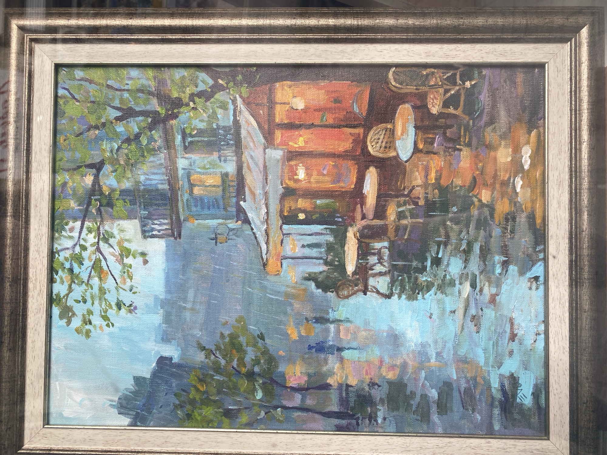 The image is a framed oil painting depicting a waterside scene, potentially a canal or river, in the city of Varna, Bulgaria. A building with orange-colored walls and multiple drawers (possibly a storage structure or workshop) is reflected in the water, along with nearby trees and foliage. A small section of what appears to be a wooden fence or gate stands partially open on the left side. The water’s surface reflects the light and colors of the environment, creating a shimmering effect with visible brushstrokes that add to the painting's impressionistic style.