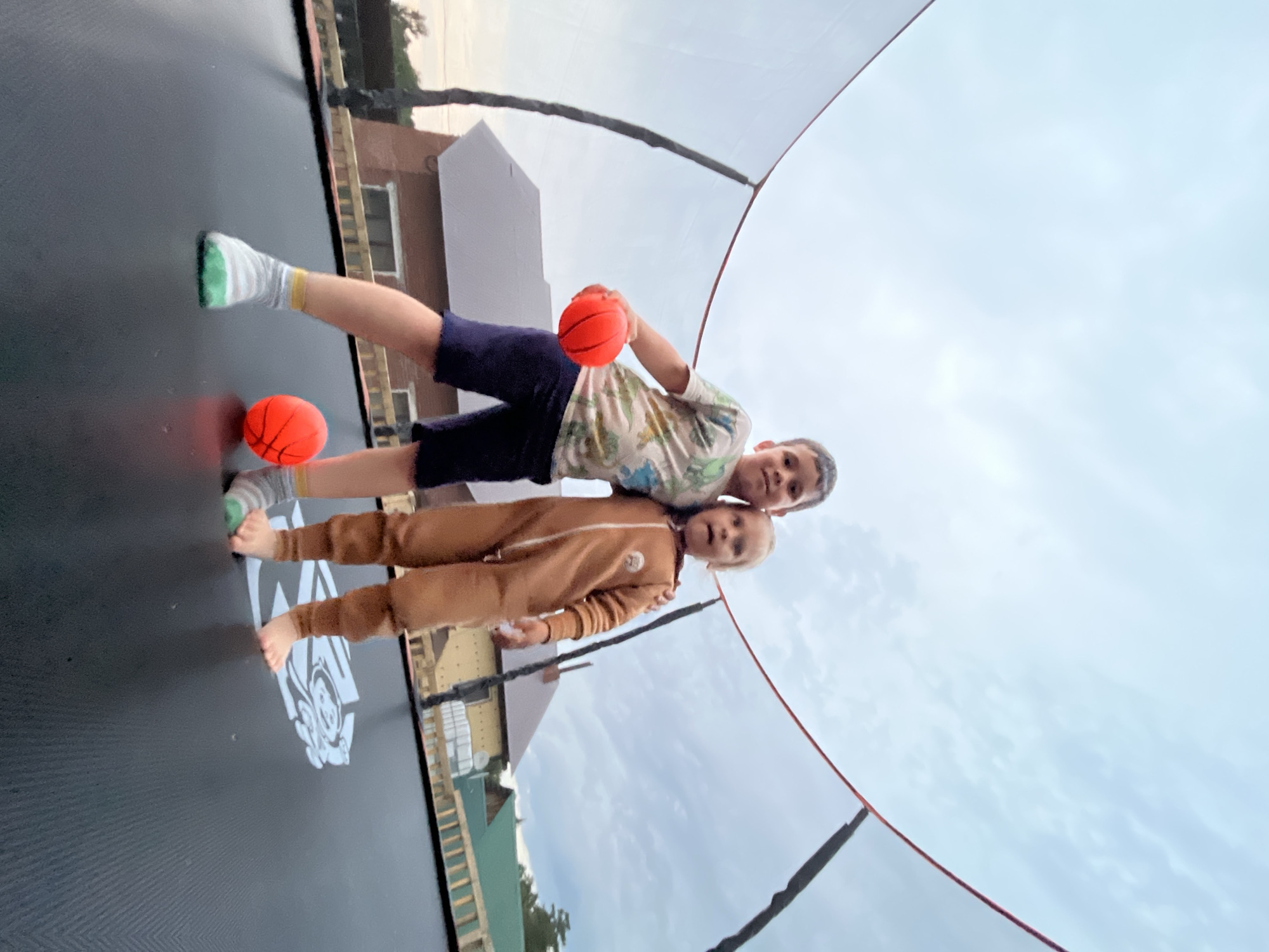 Two children, a boy and a younger girl, are standing on a black trampoline under an overcast sky. The boy, on the left, is wearing a patterned t-shirt and navy shorts, and is holding a red ball in his right hand. The girl, on the right, is wearing a tan hooded jumpsuit. Both children are looking towards the camera. There are two red balls on the trampoline. Behind the trampoline, houses with brick and green roofs are visible, suggesting a suburban or rural setting. The trampoline is surrounded by a safety net. The image appears to have been taken outdoors in the city of Chaikovsky, Russia.