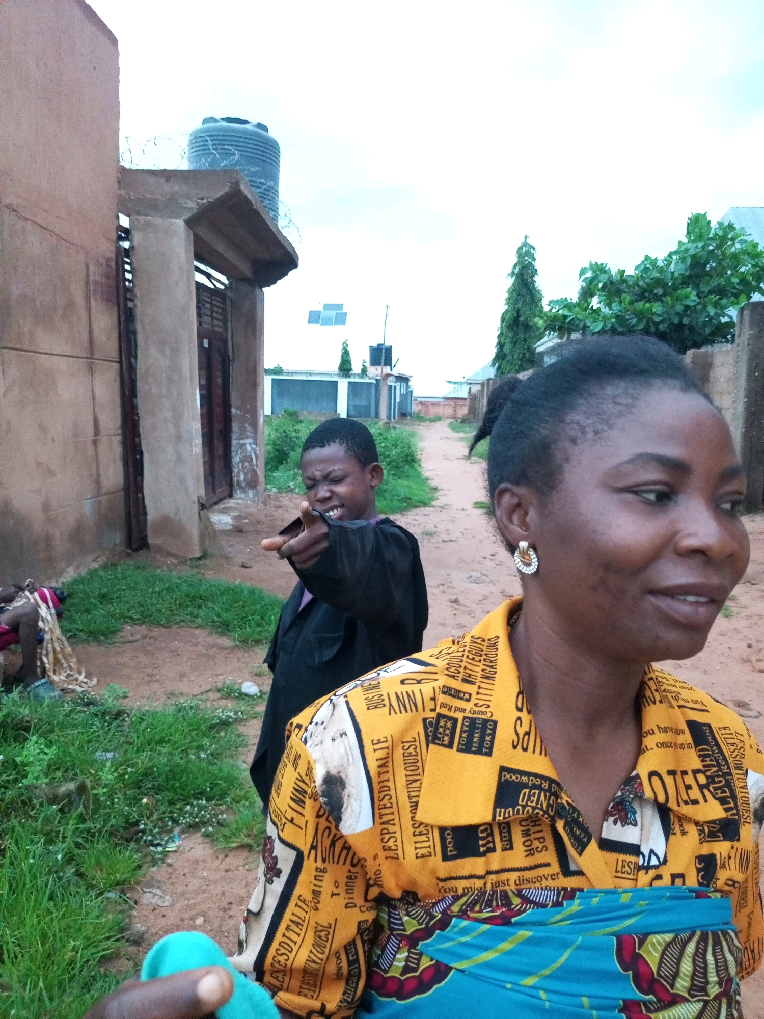 The photograph is taken outdoors in what appears to be a residential area in Tempure, Nigeria. In the foreground, a woman with dark skin and her hair pulled back in a bun is looking to her right, smiling. She is wearing a bright yellow shirt with a repeating pattern of text and images, and a blue and green patterned cloth is wrapped around her waist. A young person with dark skin is in the mid-ground, pointing to the left with an expression of mock anger. They are wearing a black garment. In the background, a dirt path leads away from the viewer. To the left, there is a building with a pinkish-brown exterior and a dark wooden door. Above this building, a large black water tank is visible, surrounded by barbed wire. Further in the background, there are other buildings and lush green vegetation. The sky is overcast.