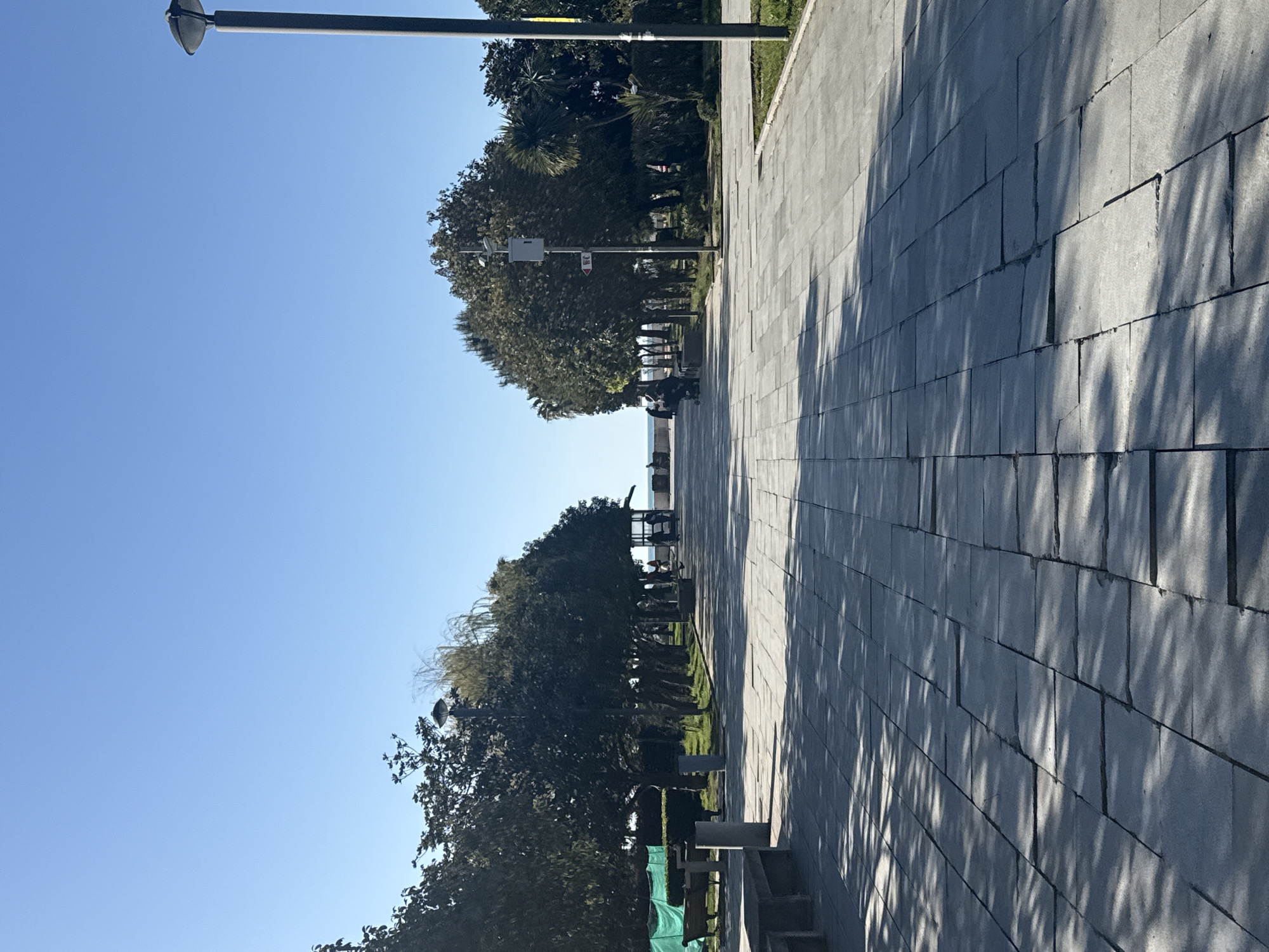 A sun-drenched outdoor scene in Batumi, Georgia, displays a wide paved promenade bordering a lush green park on a clear, bright day. The foreground features a light gray tiled pathway stretching from the lower right towards the distant horizon, with long, distinct shadows of trees cast across its surface, indicating strong sunlight, likely in the morning or late afternoon.
To the left of the pathway, a continuous line of dense green trees and shrubs forms a verdant boundary, separating the promenade from a grassy area and additional trees in the mid-ground. Several black streetlights are spaced along the path, with one prominent in the upper central portion of the image, featuring a round luminaire.
In the middle distance, small figures are visible, some appearing to be seated on benches, others possibly walking, enjoying the outdoor setting. Beyond the park and trees, a glimpse of a body of water, likely the Black Sea, can be seen, along with faint outlines of distant structures.
A utility pole in the mid-ground holds a white rectangular sign with indecipherable red text. Near the bottom left, a portion of a green tarp is visible, possibly covering equipment or indicating maintenance. The overall atmosphere is calm and pleasant under a bright blue, cloudless sky.