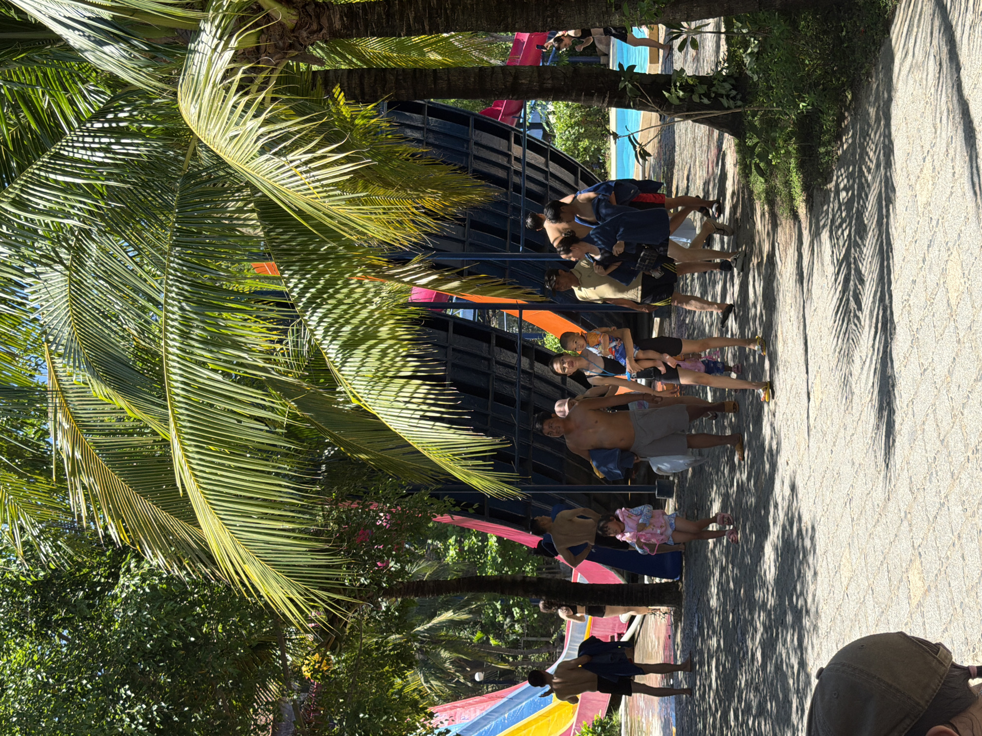 In Tay Nha Trang Ward, Vietnam, on a sunny day, a group of people are gathered at a water park near a large, multi-colored water slide complex. The foreground is dominated by palm trees casting dappled shadows on a paved walkway.
To the left, a man wearing black shorts and no shirt stands facing away from the camera, holding a dark blue towel. Next to him, a child in a pink life vest and patterned shorts walks towards the right. Behind them, a portion of a water slide with red, yellow, and blue sections is visible.
In the center, a family is walking. A man in gray shorts and no shirt is carrying a child. Beside him, a woman in a blue dress holds the child, and another woman in a dark blue outfit is walking beside them.
Further to the right, more individuals are seen walking, some holding towels. A bright blue swimming pool is visible in the background, with a person in a black swimsuit standing by its edge. The environment is lush with green trees and foliage, suggesting a tropical setting. The people appear to be enjoying a day at the water park.
