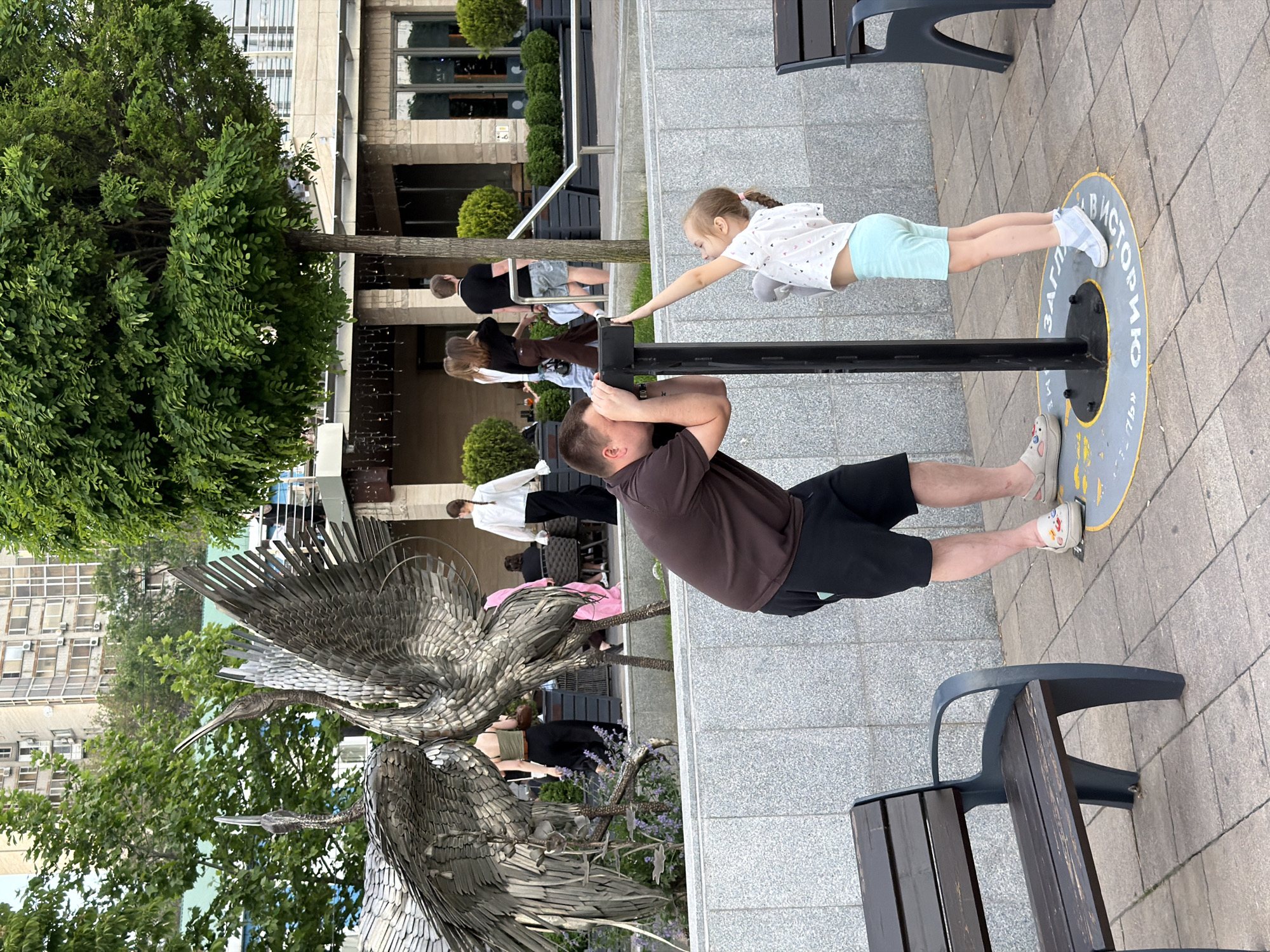 The image depicts a father and daughter interacting with a public art installation in Rostov-on-Don, Russia. The father, wearing a brown shirt and black shorts, is supporting himself on a metal bar, apparently using a camera to photograph or video his daughter. The daughter, a young girl with a ponytail, is positioned on the other side of the bar, extending her arm as if performing a balancing act. The installation includes a circular base with the inscription "заглядываю в историю" ("looking into history") visible on the ground where the father and daughter are standing. The other part of the installation is a sculpture of two large birds, likely cranes, made from metal. Several other people are in the background, including individuals dining at an outdoor cafe, and a person taking photos of the sculpture. The background also features a building with windows and greenery. Benches are placed on the paved ground in the foreground, indicating this is a public space.