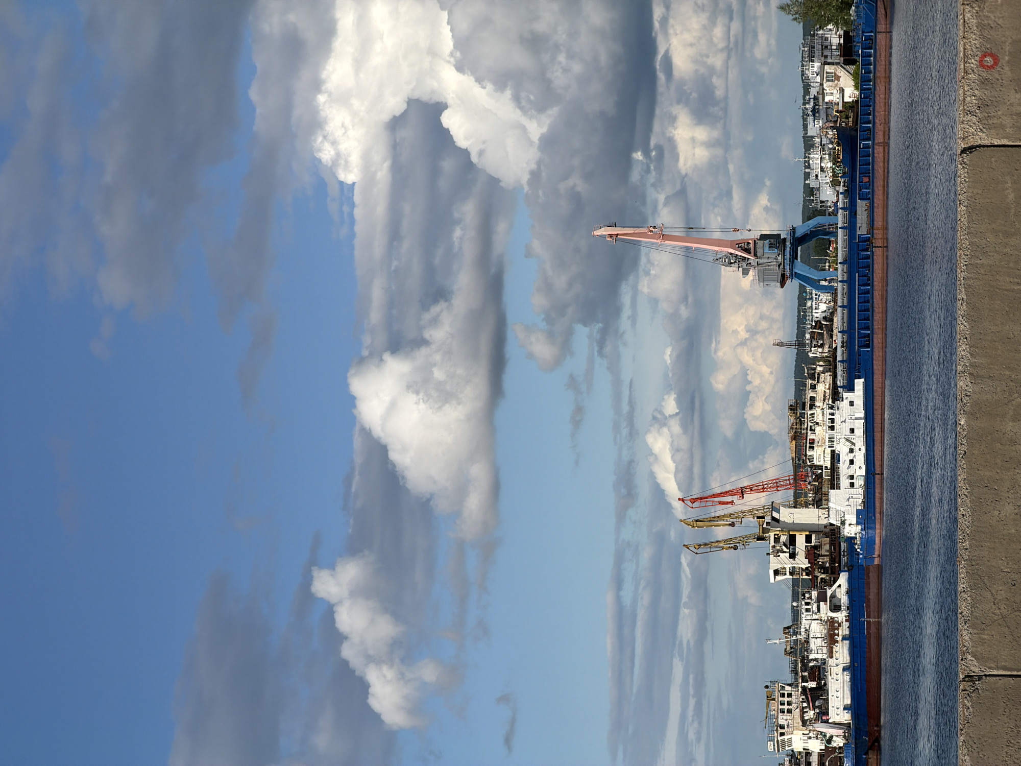 This is an outdoor scene, likely a port or dock area. The image features a sky with white and gray clouds against a blue backdrop. The foreground contains a concrete structure with water visible behind it. Cranes in shades of orange and blue are positioned near ships that are white with orange and blue features. The image is rotated, making the sky appear on the left. The location is Chaikovsky, Russia.