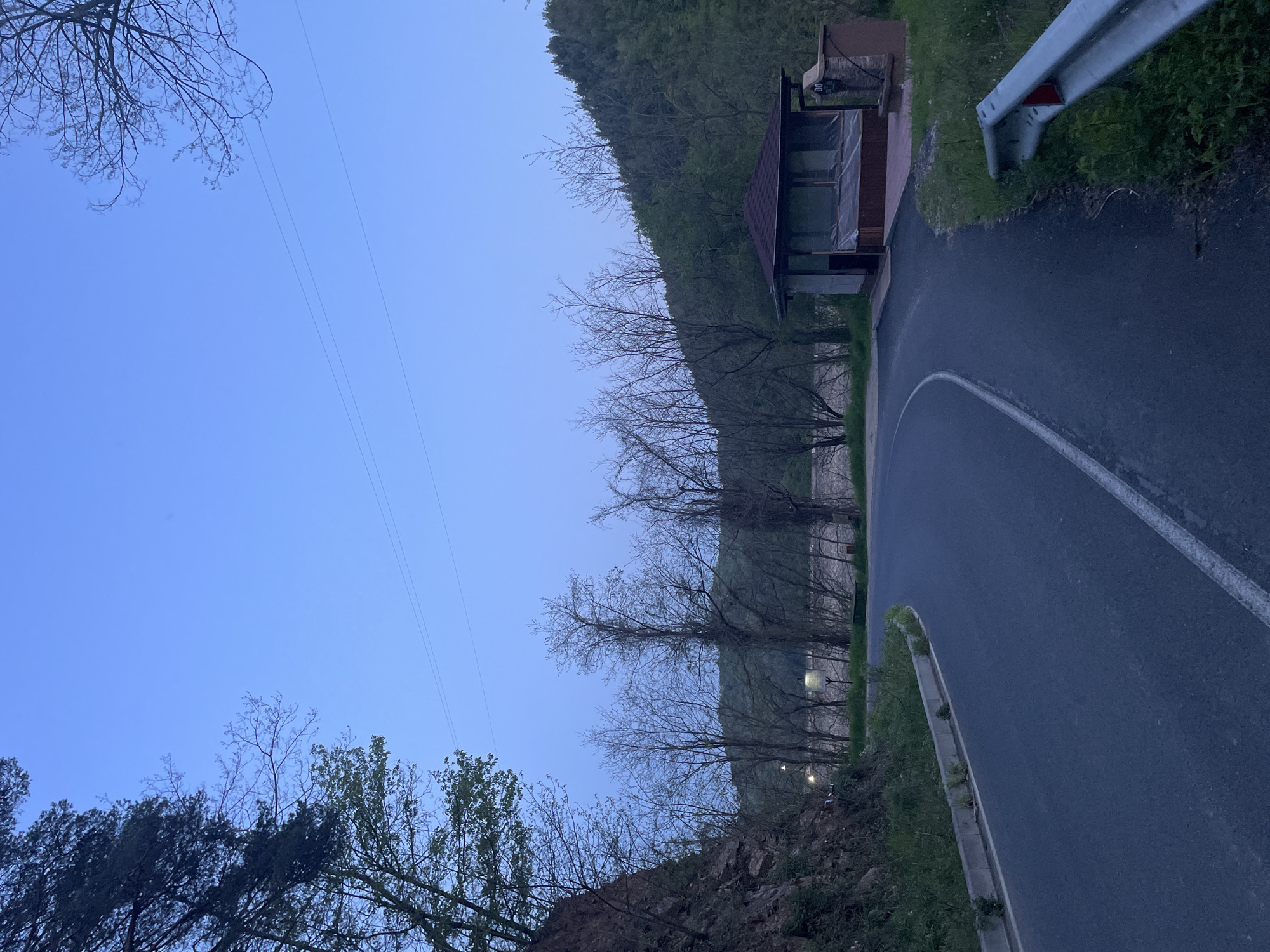 A road curves to the right through a wooded area, with a guardrail on the right side of the road. A small structure with a roof and windows sits alongside the road. Trees line the left side of the road, with a hillside visible in the background. The sky is blue, suggesting early morning or dusk. The location appears to be a rural area near Zlatograd, Bulgaria, based on the presence of characteristic regional vegetation and architecture. No people are visible.