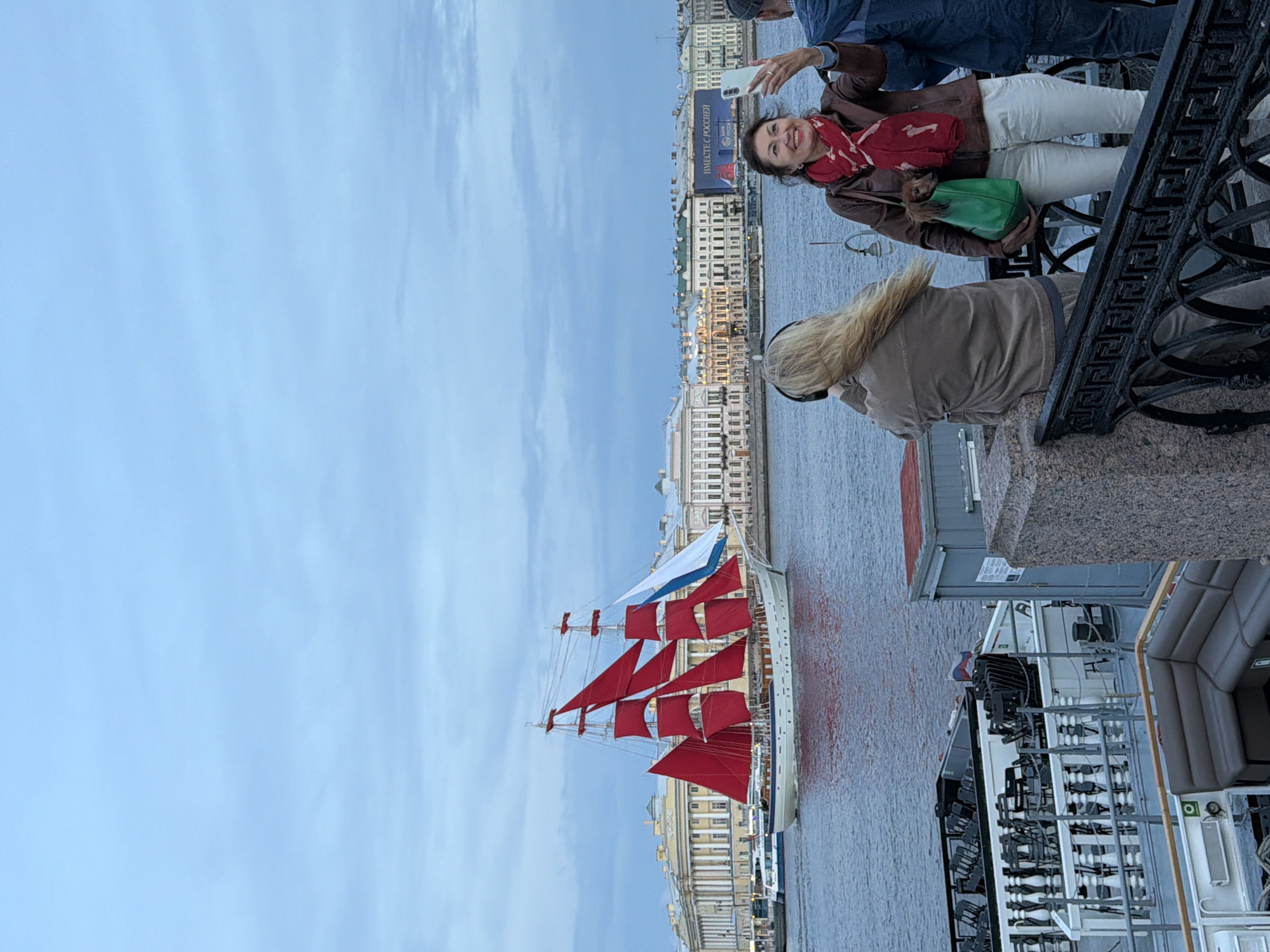 A three-masted schooner with bright red sails and a single white sail is sailing on the Neva River in Saint Petersburg, Russia. The ship is passing by a large building complex with ornate architecture that lines the riverbank. In the foreground, on a promenade, two women and a man are visible. One woman, with blonde hair and wearing headphones, is looking away from the camera. The other woman, with dark hair and wearing a red scarf, is taking a selfie with her smartphone and holding a green bag with a small dog peeking out. The man, wearing a dark hat and jacket, is standing next to her, partially visible. The sky is overcast with pale blue and grey clouds. The water of the river is a deep blue. There are also parts of a moored boat with seating and railings in the foreground.