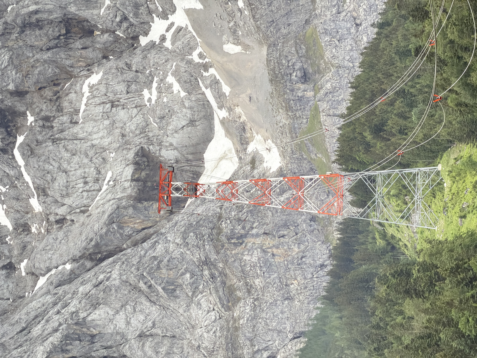 A tall, red and white lattice tower stands in the foreground, supporting cables for a cable car. The tower is situated on a grassy slope covered with trees. Behind the tower, a massive, grey, rocky mountain rises, with patches of snow and scree visible on its face. The scene is set in the Bavarian Alps, near Grainau, Germany, likely a station for the Zugspitzbahn cable car. No people or animals are visible in the image.