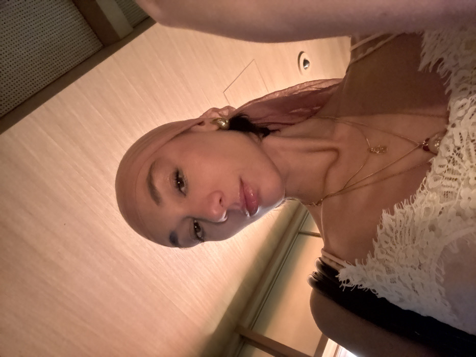 A young adult female is depicted in a close-up, low-angle selfie. She wears a sheer, light peach-colored headscarf wrapped around her head, covering her hair. Her face is lightly made up, featuring glossy lips, visible eyelashes, and defined eyebrows. A single gold-toned hoop earring is visible on her right ear. She is wearing multiple layered gold-toned necklaces; one is a thin chain with a small, elongated pendant, and another is a longer chain with a larger, possibly red and gold, ornate pendant. Her attire includes a light-colored, likely cream, top with intricate lace detailing visible across the bust and a shoulder strap. Dark hair is visible at the nape of her neck on the right side, emerging from beneath the headscarf. The indoor background primarily shows a light-colored, possibly wooden, paneled ceiling illuminated by warm, ambient overhead lighting that casts a reddish-orange hue. A small, recessed fixture or vent is partially visible on the upper right side of the ceiling. There are no discernible visual cues within the image to definitively identify the location as Paris, France.