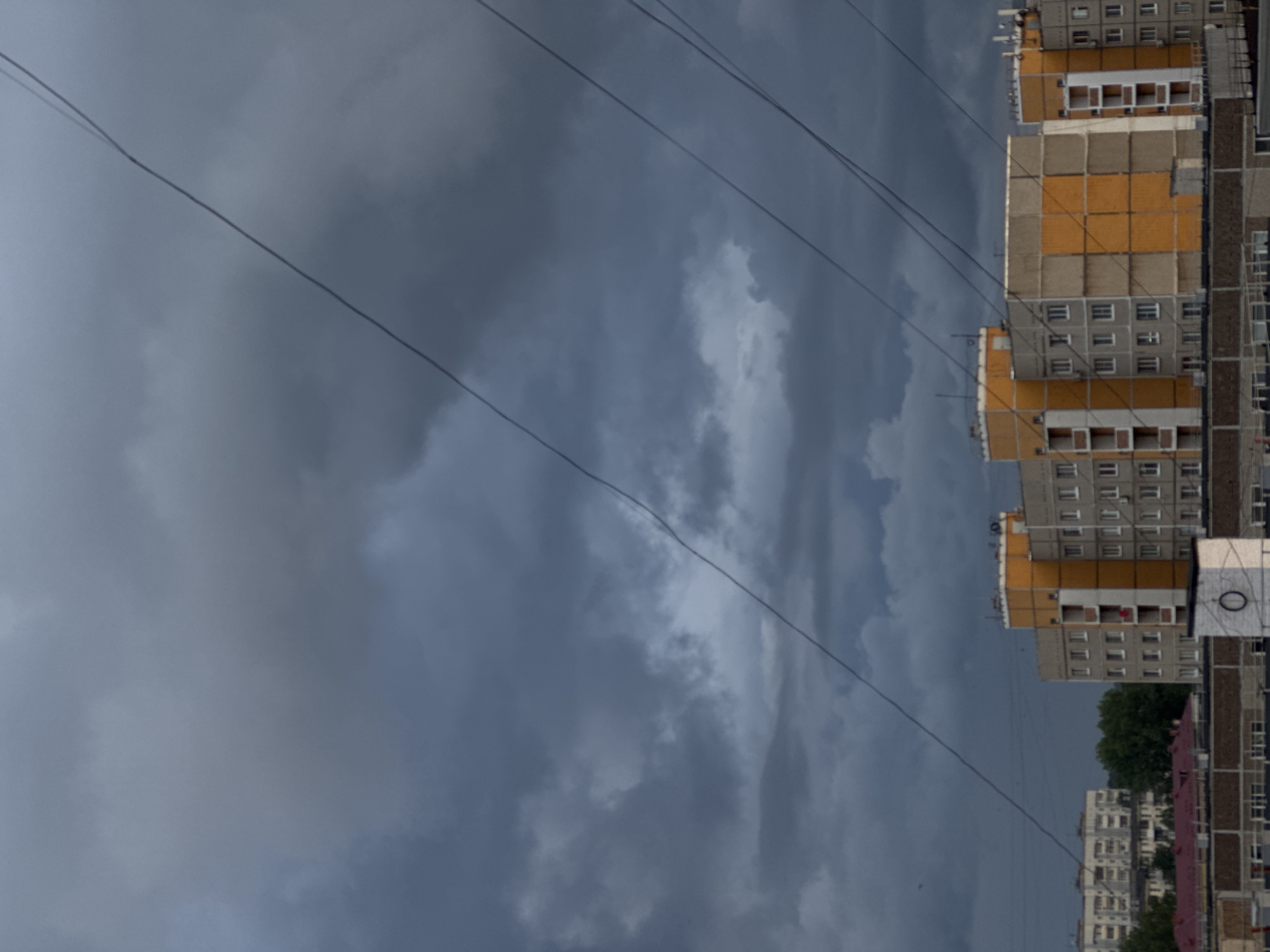 The image captures an urban landscape in Chaikovsky, Russia, dominated by a dramatic, overcast sky. Heavy, dark grey clouds fill most of the frame, suggesting an impending storm or recent rain, contributing a moody and diffused light to the scene. Several dark power lines or communication cables stretch diagonally across the sky, adding linear elements that cut through the expansive cloud formations.
On the right side, multiple multi-story residential buildings, likely apartment blocks, rise into view. These structures feature facades with alternating sections of grey concrete and brighter yellow or orange panels, characteristic of Soviet-era or post-Soviet architecture. Numerous windows are visible across their surfaces. A small cluster of green trees is nestled between and behind some of the buildings in the lower right portion of the frame.
No people or specific activities are visible, presenting a static moment of urban infrastructure under a powerful natural backdrop. The low, diffused light indicates either a dark, heavily clouded day or possibly late afternoon/early evening. The overall mood conveyed is somber or anticipatory due to the dramatic sky. A very faint, indistinct white square-like structure with a dark circular element, possibly resembling a "0" or an "O," can be barely discerned on one of the buildings on the far right.