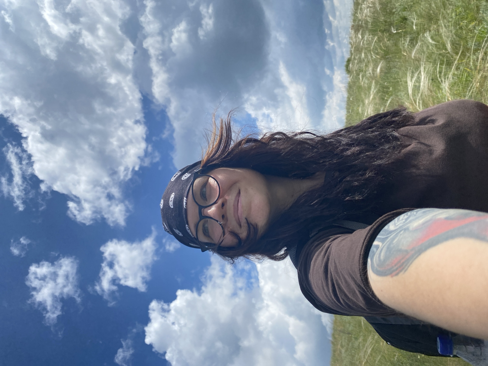 A woman with dark wavy hair, wearing a brown t-shirt and a black bandana with white patterns, and round glasses, is taking a selfie outdoors. She has a tattoo on her left arm. She is in a grassy field with tall, feathery grass swaying in the wind. The sky is blue with fluffy white and grey clouds. The location is Yuldybaevo, Russia.
