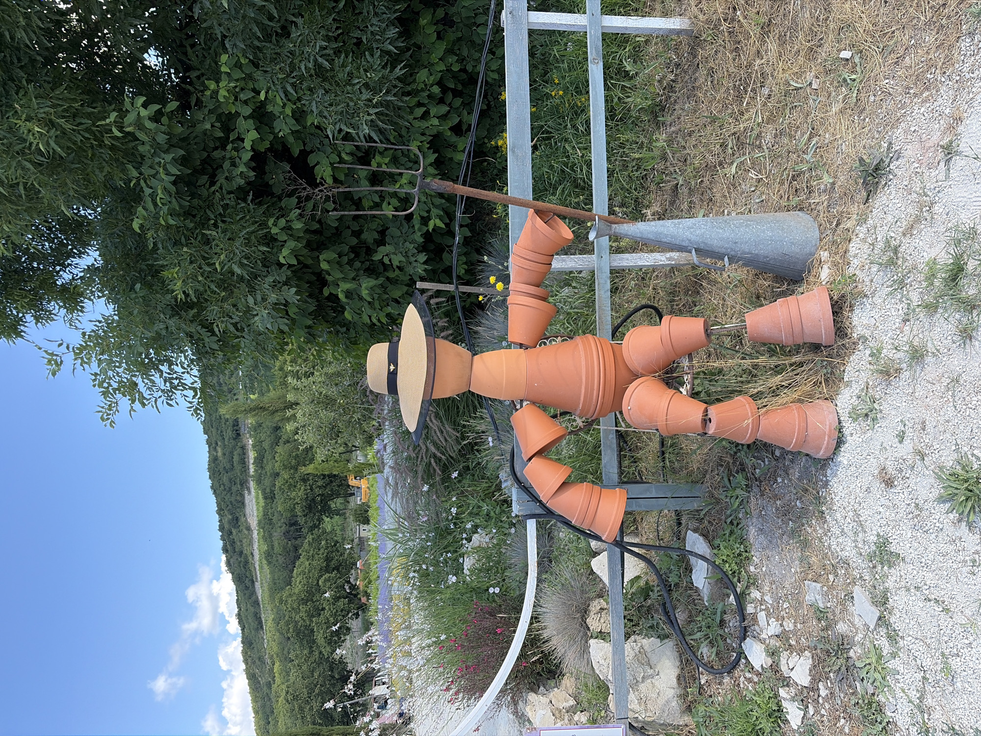The image shows a scarecrow constructed from terra cotta pots, standing in a garden setting. The scarecrow is wearing a straw hat adorned with a bee emblem. It is holding a garden fork and a metal funnel. The background features lush greenery, flowering plants, and a hilly landscape under a clear blue sky with scattered clouds. The ground is covered with gravel and dry grass. The scene is set in Semogorskiy, Russia.