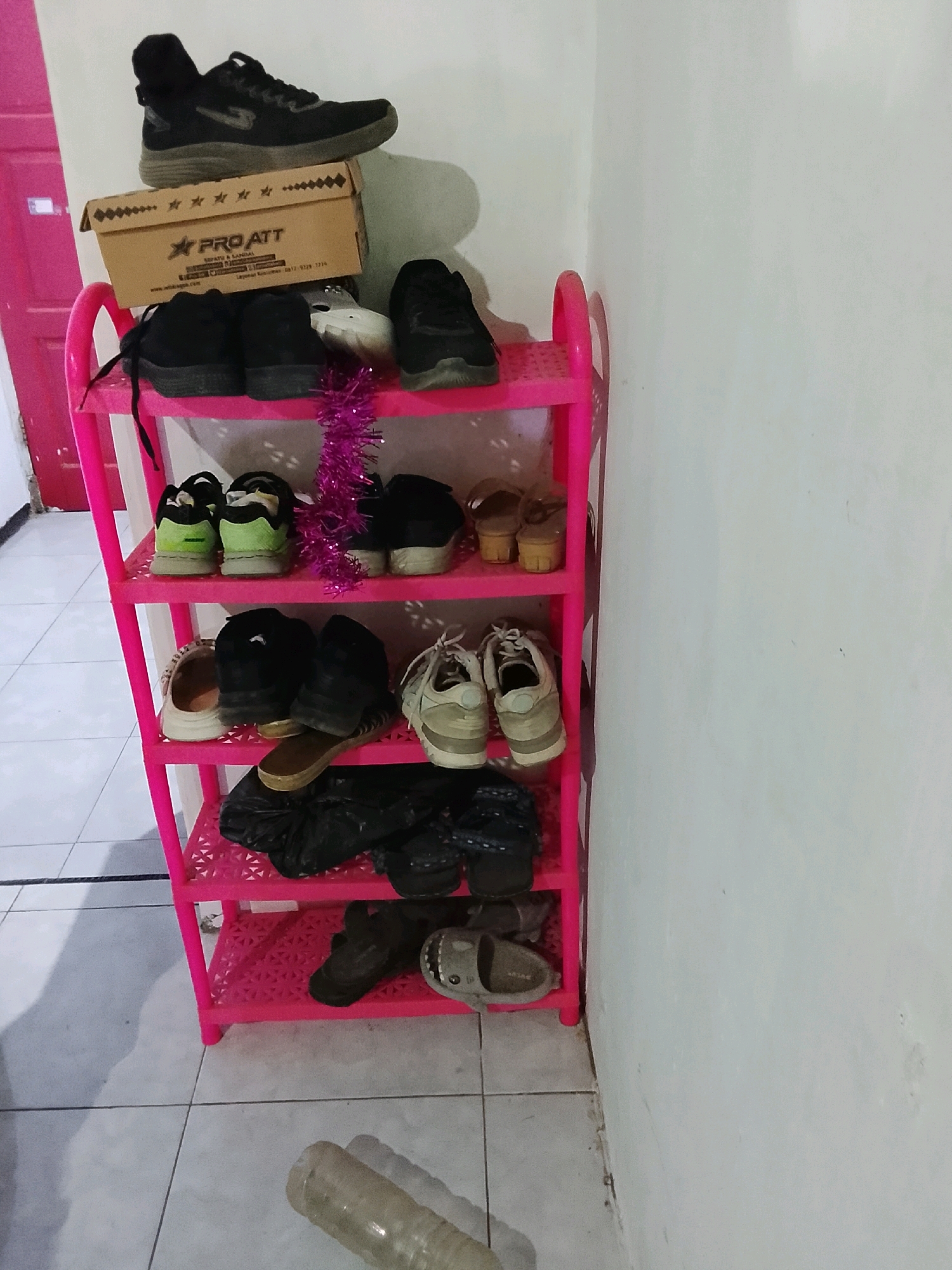 An indoor scene in a building in Ujungrusi, Indonesia, features a pink plastic shoe rack filled with various footwear. The rack is positioned against a white wall. On the top shelf, a pair of black high-top sneakers rests on a cardboard box labeled "PRO ATT". Below this, the second shelf holds a pair of black sneakers and another pair of black shoes. The third shelf displays two pairs of shoes: one pair of light-colored sneakers and another pair of black shoes. The fourth shelf contains a pair of beige clogs and a pair of black sneakers. The bottom shelf has a black plastic bag and a pair of gray sandals. A clear plastic bottle lies on the tiled floor in the foreground. The overall impression is one of a functional storage area.