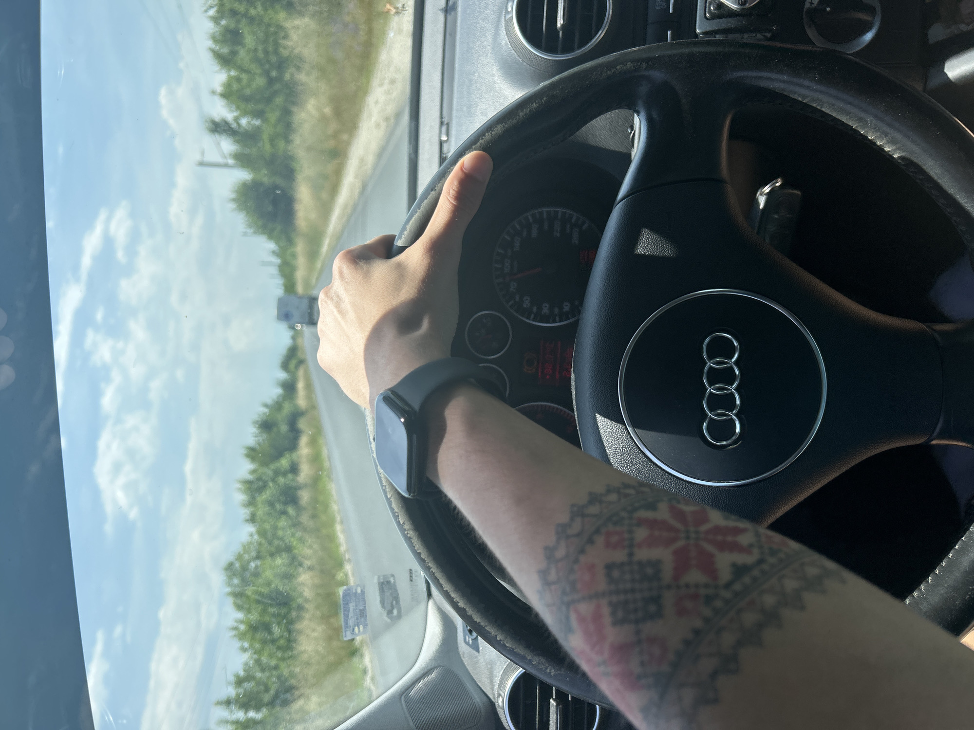The image captures a first-person perspective from inside a car, showing a driver's left arm and hand gripping the steering wheel. The driver is wearing a dark-colored smartwatch on their wrist, and a prominent, colorful tattoo featuring a geometric, folk-art pattern with red elements is visible on their forearm.
The steering wheel is black, with the distinctive silver Audi logo (four interlocking rings) centered on the airbag cover. The instrument cluster is partially visible through the top-left section of the steering wheel, displaying a speedometer with markings from 0 to 260. Some dashboard warning lights are also faintly discernible at the bottom of the cluster. The car's interior appears to have black and gray tones, with an air vent visible on the right side of the dashboard.
Through the front windshield, a bright, sunny day is visible with a clear blue sky and scattered white clouds. A paved road stretches into the distance, flanked by lush green trees and vegetation on both sides, suggesting a rural or semi-rural setting. The strong sunlight indicates midday or early afternoon. The scene depicts the activity of driving a vehicle on a road. Given the provided context, the location is Dalgo Pole, Bulgaria.