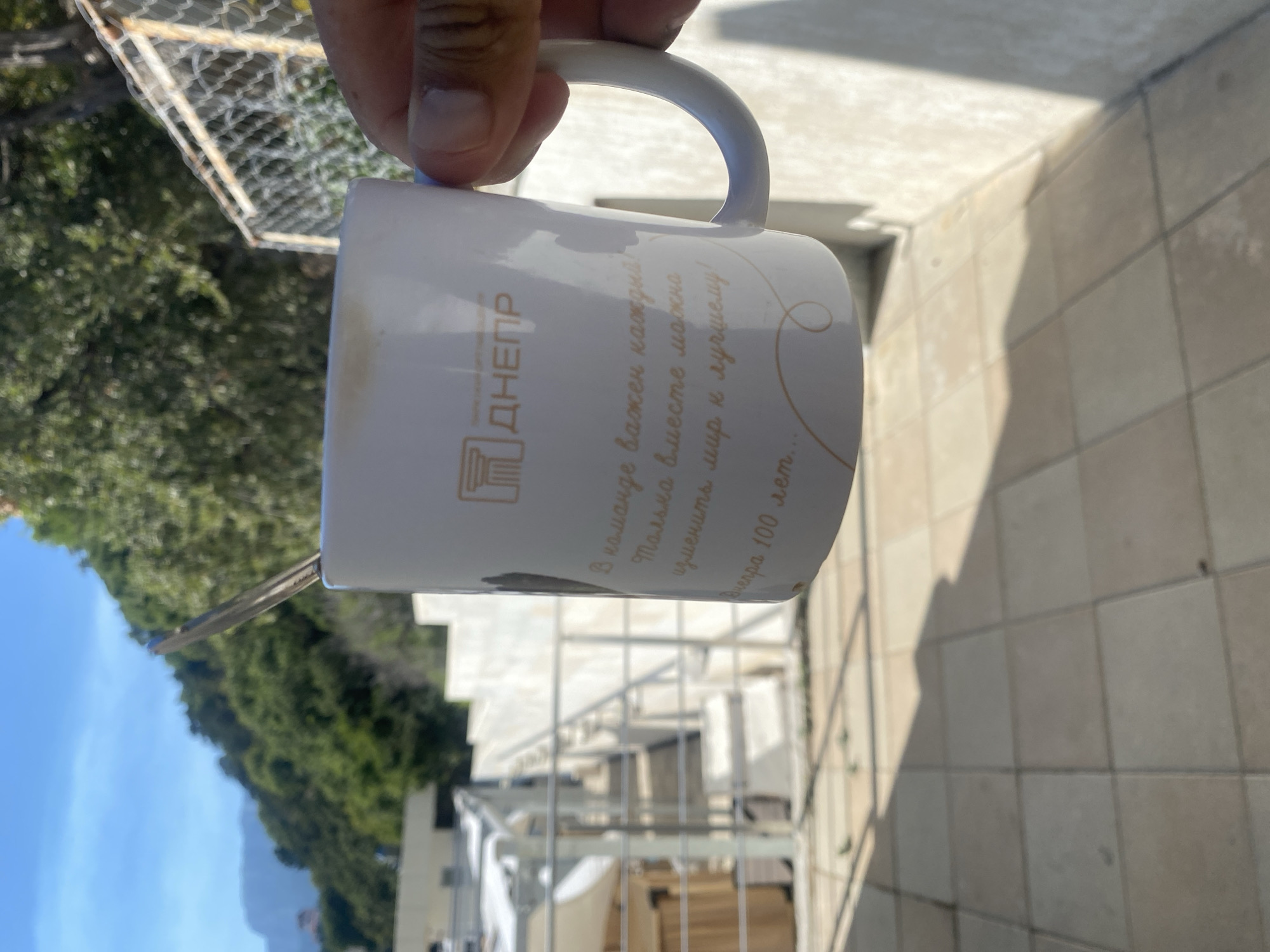 A person is holding a white mug with a spoon in it. The mug has a logo with the Cyrillic letters "ДНЕПР" and some text that appears to be in Russian. The background shows a tiled patio, a metal fence, and a hill with trees. The location is the village of Gaspra, Yalta Municipality, Crimea, Ukraine. The sky is blue with some clouds, and the sun is shining.
