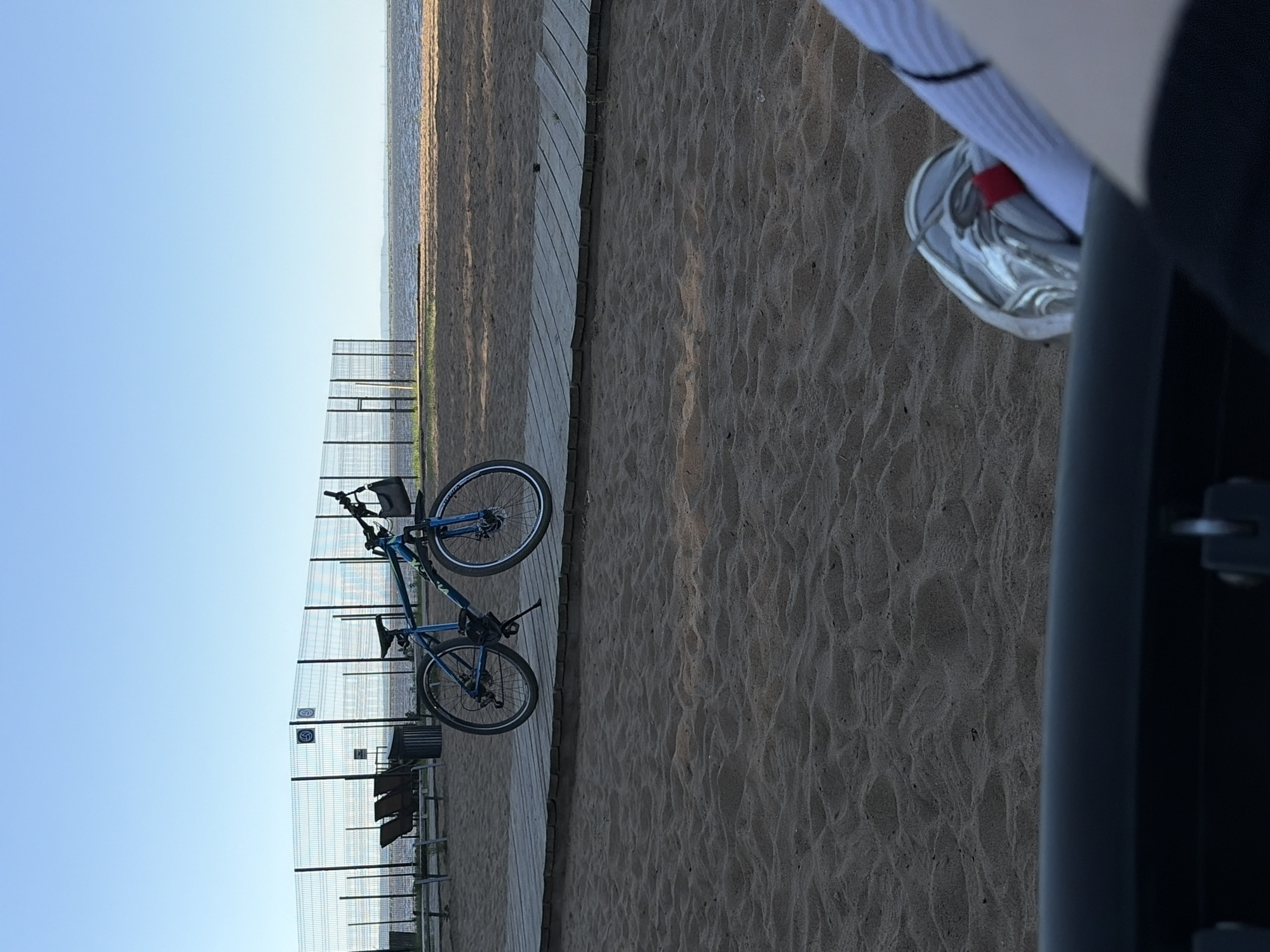 The image depicts a beach scene with a bicycle leaning against a fence in the background. A wooden walkway runs parallel to the sandy beach. In the foreground, part of a chair and a person's leg with a sock and silver shoe are visible. The sky is clear and blue, suggesting a sunny day. The scene is likely at a beach in the city of Unknown, Unknown.