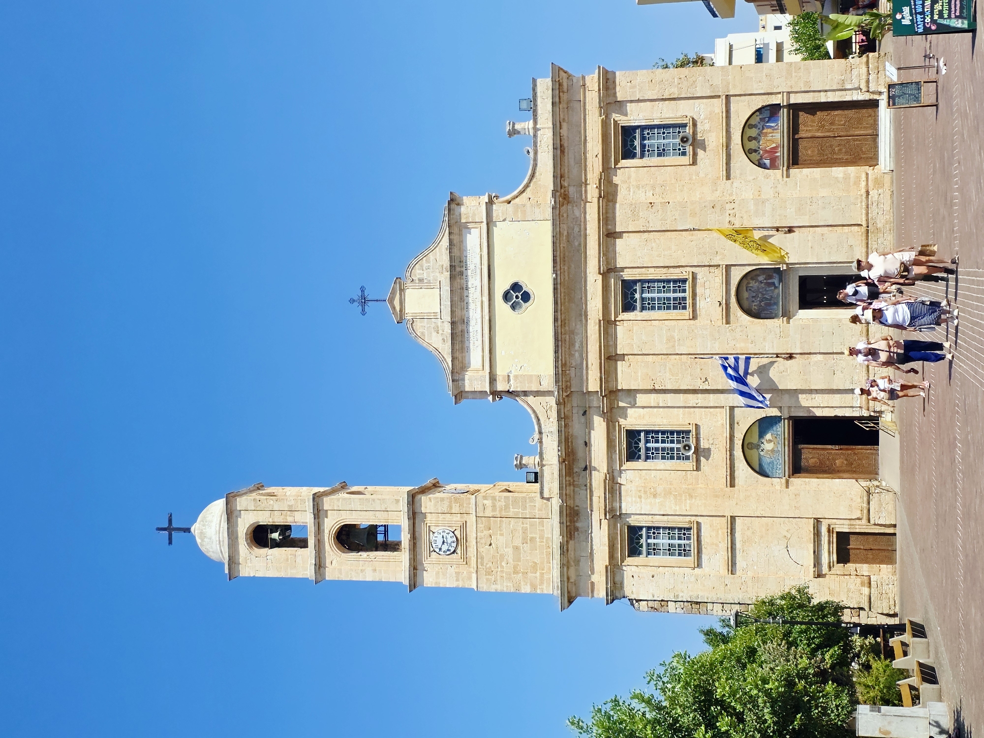 This is a vertical, eye-level, full shot of the Church of Agios Nikolaos in Chania, Greece, on a clear, sunny day. The church is made of light-colored stone and has a tall bell tower with a clock. There are two large wooden doors on the front facade, with religious paintings above them. Two flags, one Greek and one yellow, are displayed on the building. Several people are walking in front of the church, some appearing to be tourists. Green trees and benches are visible on the left side of the image, and a blackboard sign for a cafe is on the right. The ground in front of the church is paved with reddish-brown bricks. The sky is a clear, bright blue.