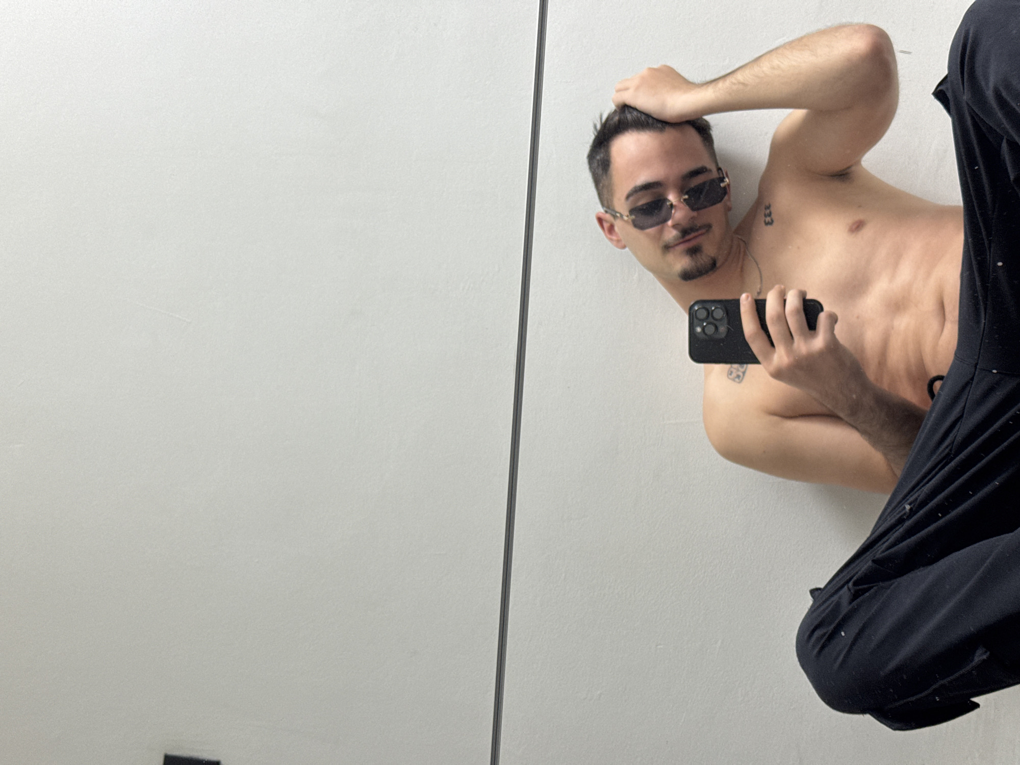 A young man is depicted in a mirror selfie, appearing shirtless and wearing dark sunglasses with gold-toned accents. He is posed against a plain, light-colored wall, which is reflected in the mirror. He holds a black smartphone in his left hand, seemingly capturing his reflection, while his right hand rests on the top of his head. He has short dark hair, a mustache, and a goatee.
The man exhibits a lean physique with visible abdominal muscles. He has a small tattoo of "333" on his right shoulder/chest area and another indistinct, smaller tattoo on his left arm. A small reddish mark or scar is visible on his right upper chest. He wears dark pants, partially visible in the lower right of the frame. The reflection shows a vertical seam running through the mirror, dividing the background. The scene is set indoors, with the location context being Sofia, Bulgaria. The lighting is bright and even.