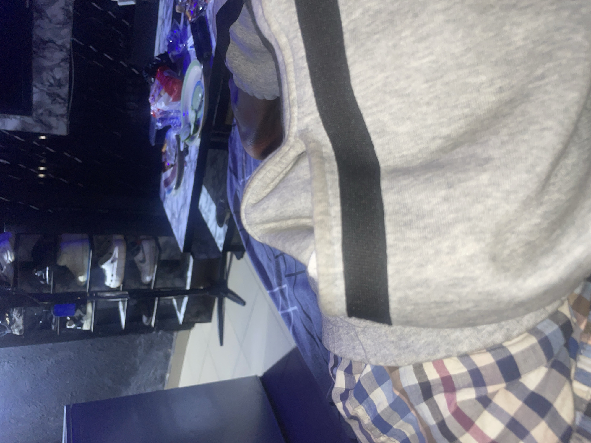 The image appears to be taken from a low angle, focusing on clothing and bedding. In the foreground, a grey sweatshirt with a black stripe is draped over what looks like a person's lap. Beneath the sweatshirt, a blue and white checkered fabric, possibly a shirt or blanket, is visible. The background shows a shoe rack filled with various sneakers and shoes, a marble-patterned table with plates and plastic-wrapped items, and a dark wall. The lighting is dim, with some blue light illuminating the left side of the frame. The scene suggests an indoor setting, possibly a bedroom or living area. The location in Rafin Par, Nigeria, is not discernible from the visual information provided in the image.