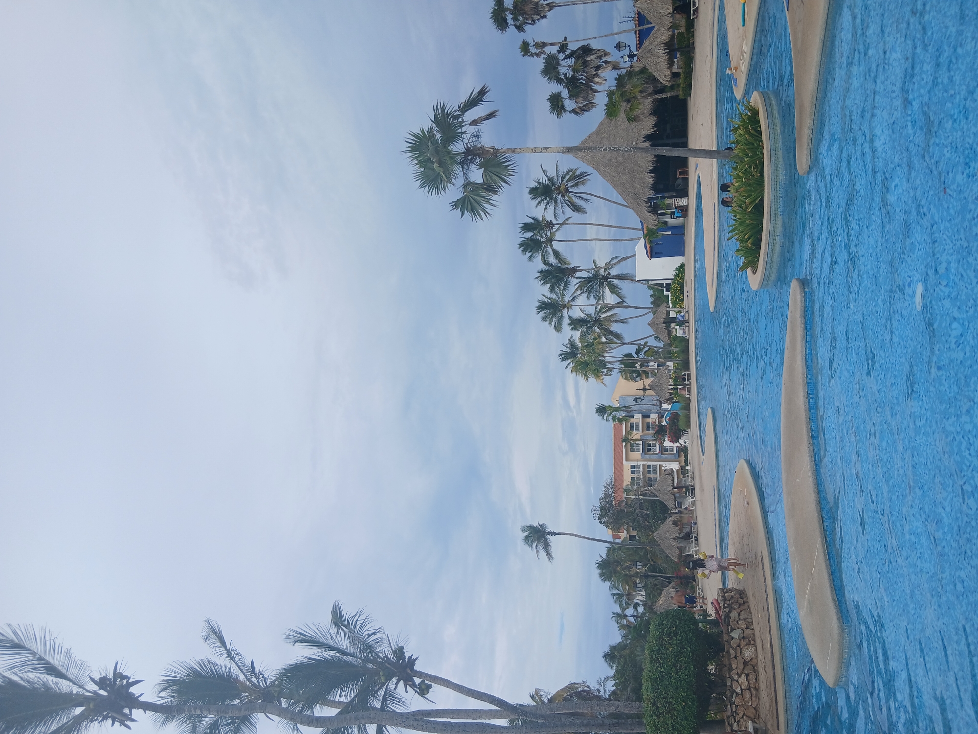 The scene depicts a resort complex in Playa El Agua, Venezuela, including palm trees, a pool area, and hotel buildings. The pool, of blue tile, curves in a unique design, seemingly artificial island-like structures. Thatched-roof structures provide shade around the pool and on a hotel roof. Palm trees populate the grounds. Individuals can be observed near the pool, though their actions and interactions are difficult to assess. The sky is mostly blue, and buildings show a few colors, including orange. The general ambience is sunny and warm.