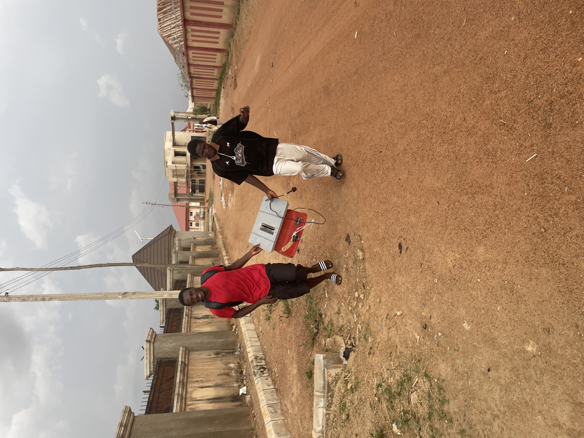 Two young men stand on a dusty, red-brown dirt road, possibly in Jos, Nigeria, based on the architectural style of the surrounding buildings. One man wears a black t-shirt with white lettering and light-colored pants. He holds a rectangular gray and red electrical box, which is connected with wires. The other man is wearing a red t-shirt and black shorts and has a backpack. Buildings and walls are visible along the roadside. Overhead electrical lines are also present. The sky is overcast.