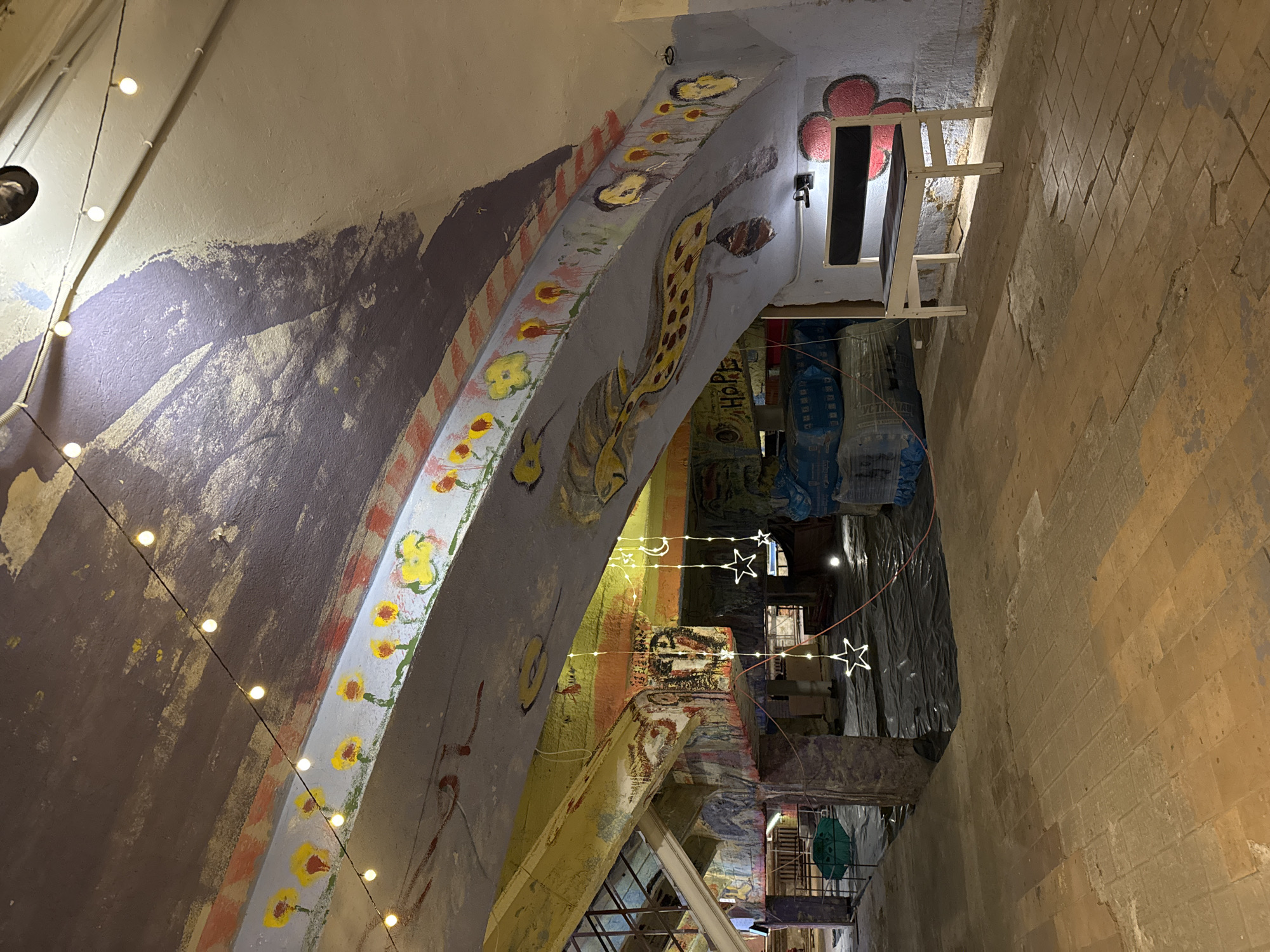The image shows the interior of the Petrikerk church in Saint Petersburg, Russia. The camera points up through a passage in the church that appears to be under construction, with exposed beams and scaffolding. The walls and ceilings of the passage are covered in graffiti and murals, including a string of lights strung along one side. There are also string lights in the distance. The floor is a brick-like pattern in shades of tan. Various objects and materials are stored in the passage.