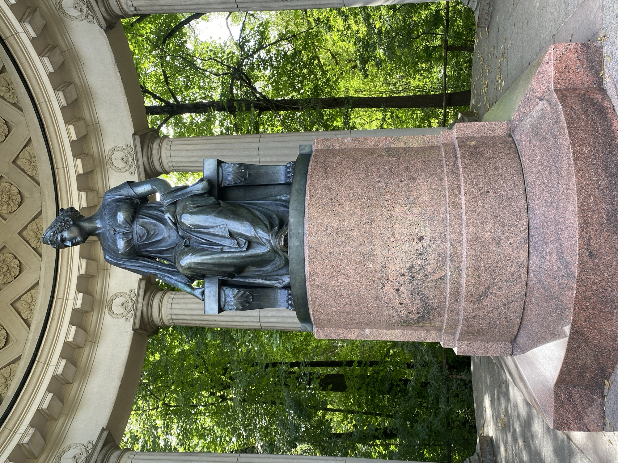 The image shows a full-body shot of a bronze statue of a seated woman, likely depicting Empress Maria Fyodorovna. The statue is situated within a neoclassical rotunda supported by fluted columns. The woman is adorned with a crown and a draped garment, and she is seated on a throne-like chair. The base of the statue is made of pink granite, with a circular, stepped design. Behind the rotunda, a lush green forest is visible, with sunlight filtering through the trees. The overall setting suggests a public park or garden, consistent with the grounds of Pavlovsk Palace in Pavlovsk, Russia, where numerous monuments and architectural features dedicated to the imperial family are located. The statue appears to be in good condition, with no visible damage. The environment is serene and natural, with the monument serving as a focal point within the landscape.
